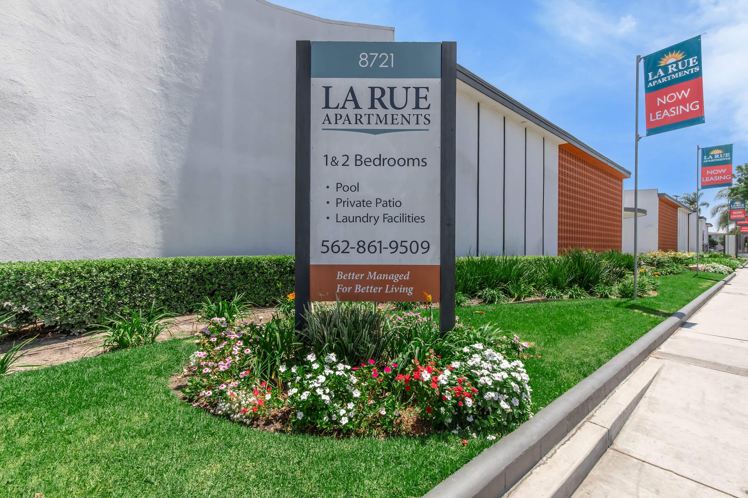 Sign for La Rue Apartments, located at 8721. Lists features including 1 & 2 bedrooms, pool, private patio, and laundry facilities. Includes contact number 562-861-9509 and mentions "Better Managed For Better Living." The sign is surrounded by landscaped flowers and greenery.
