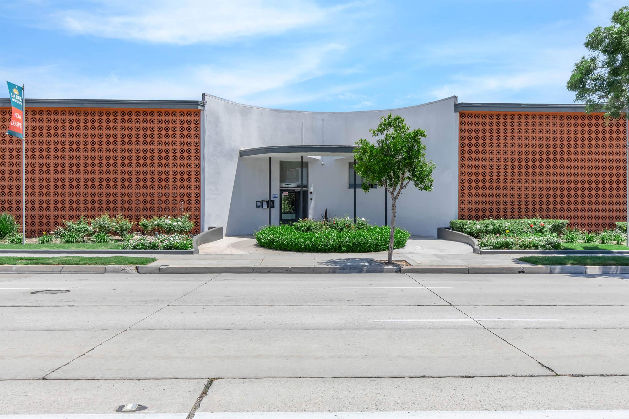 Modern building facade featuring a curved entrance with glass doors. The exterior has a geometric patterned wall in warm tones, surrounded by greenery and flower beds. A paved walkway leads up to the entrance, and the image captures a sunny day with a clear sky.