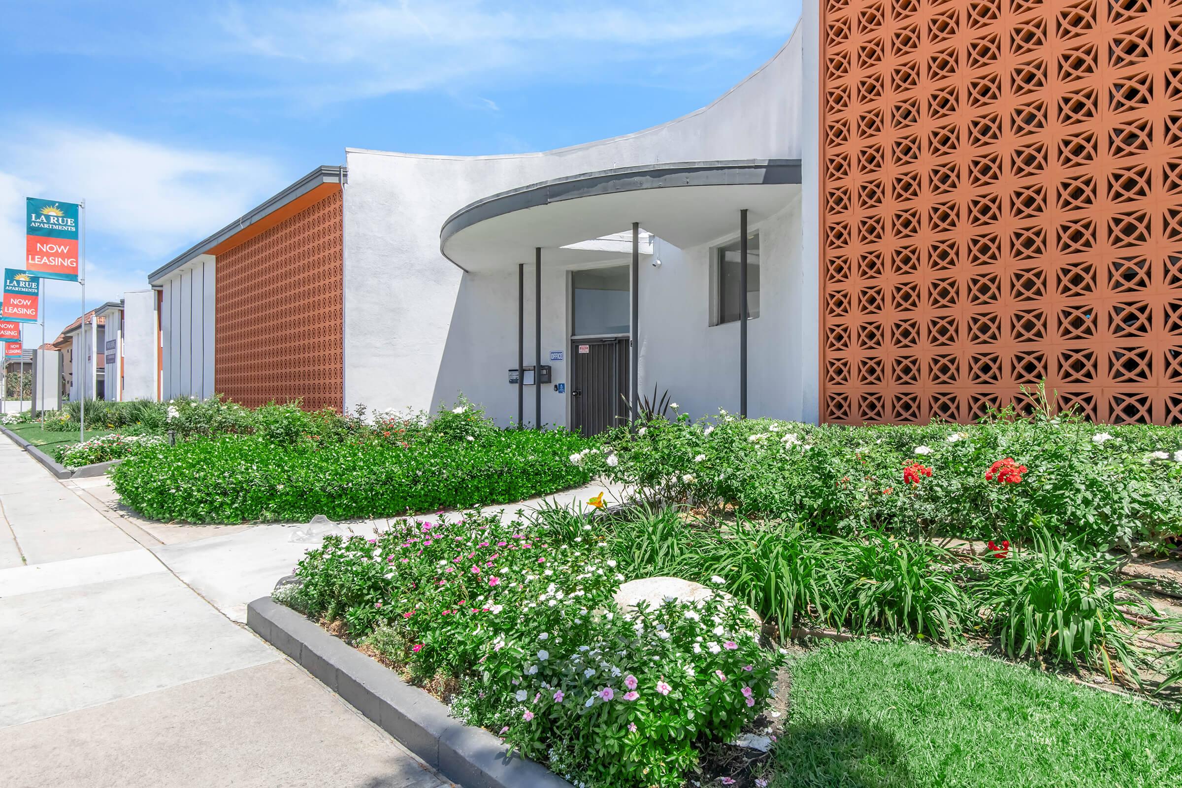 A modern building with a curved entrance and decorative brick wall, surrounded by landscaped flower beds featuring various colorful plants. The setting includes a sidewalk and signage indicating availability for leasing. Clear blue sky above enhances the attractive exterior.