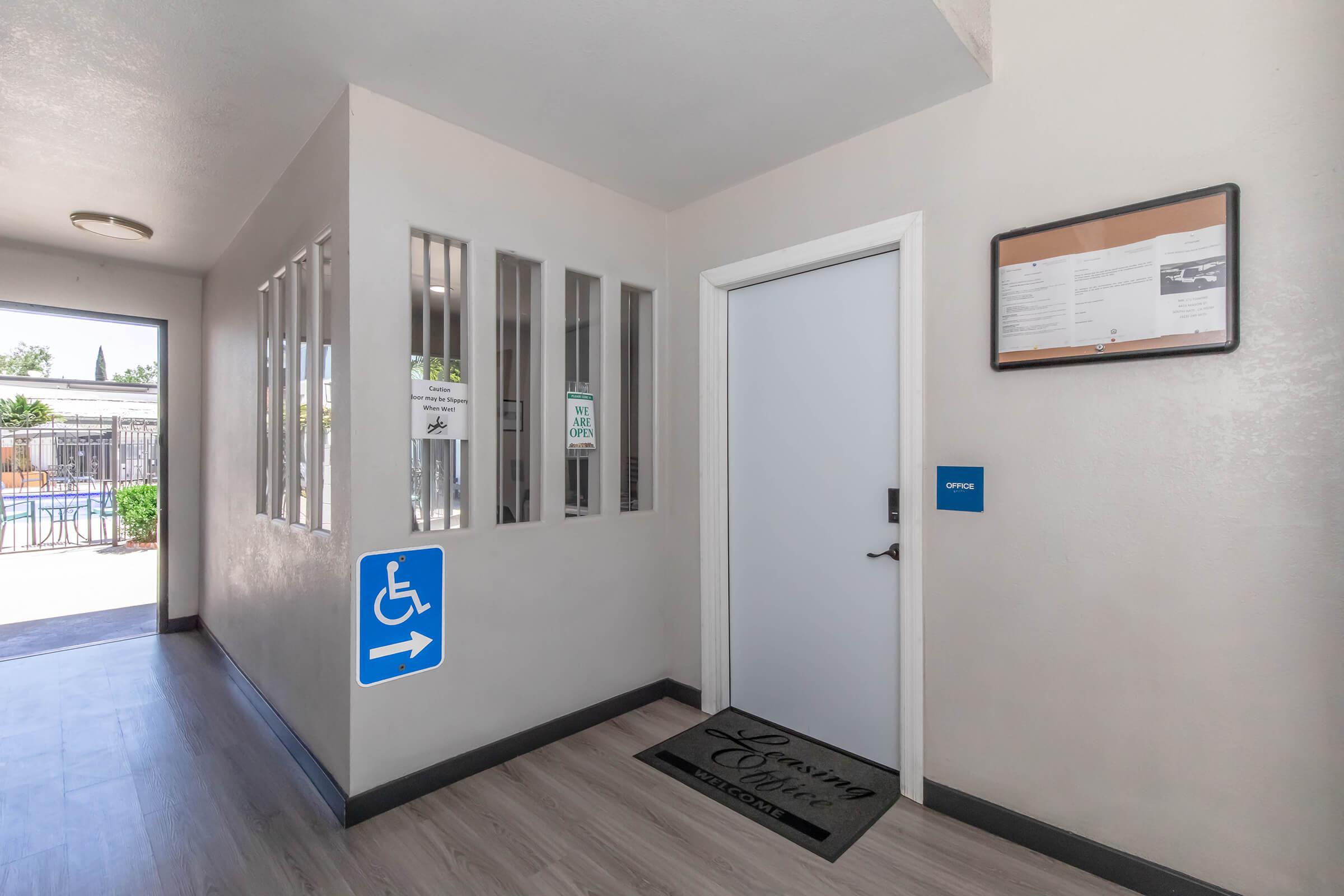 A well-lit hallway leading to an office door, featuring signs for accessible entry and office hours. There are large windows on the left, allowing natural light, and a welcome mat in front of the door. A blue arrow indicates the direction for accessible access.
