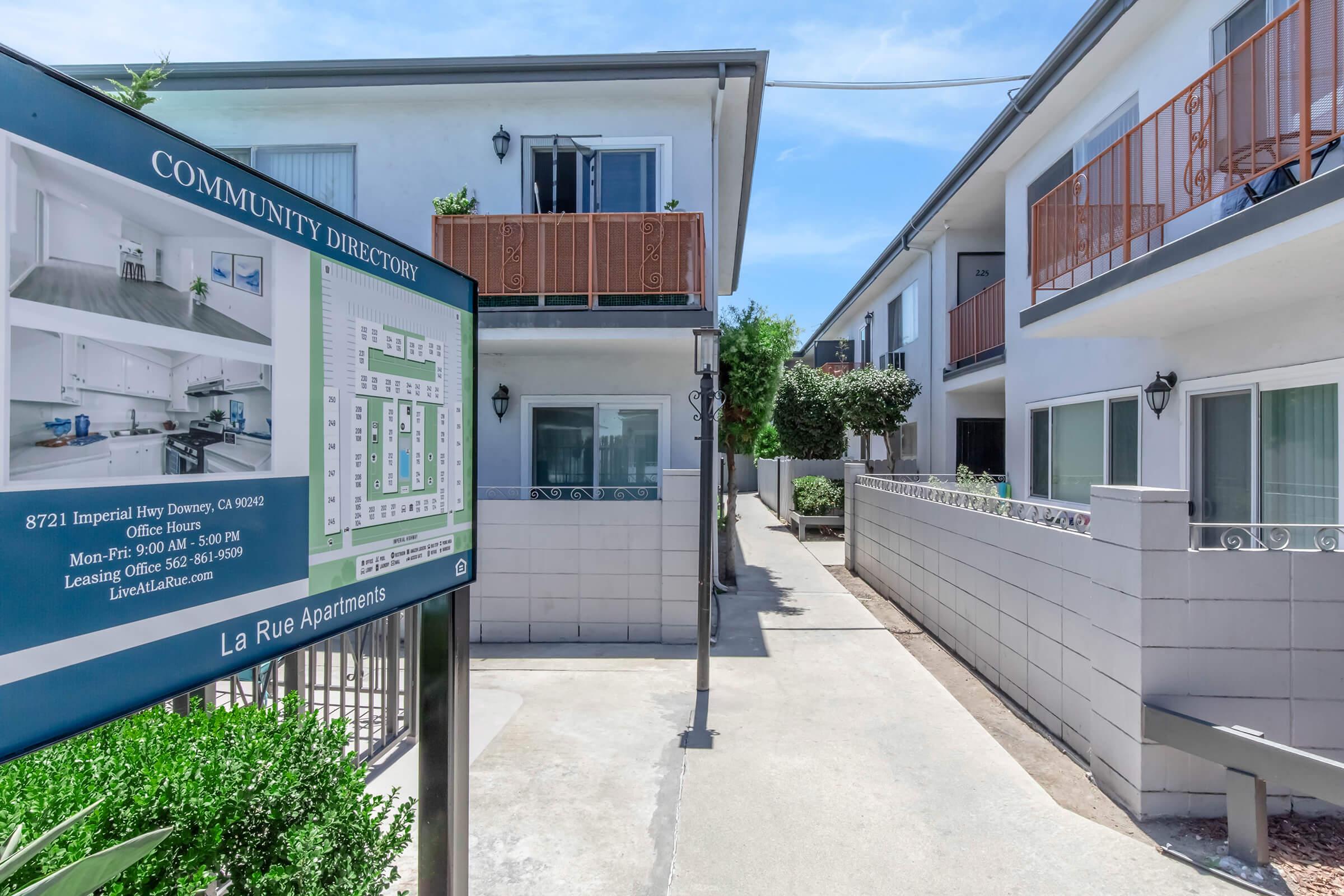 A community directory sign displaying information about La Rue Apartments, with a clear view of the apartment building and walkway leading into the complex. Green landscaping is visible, along with balconies on the upper floors and a bright, sunny atmosphere.