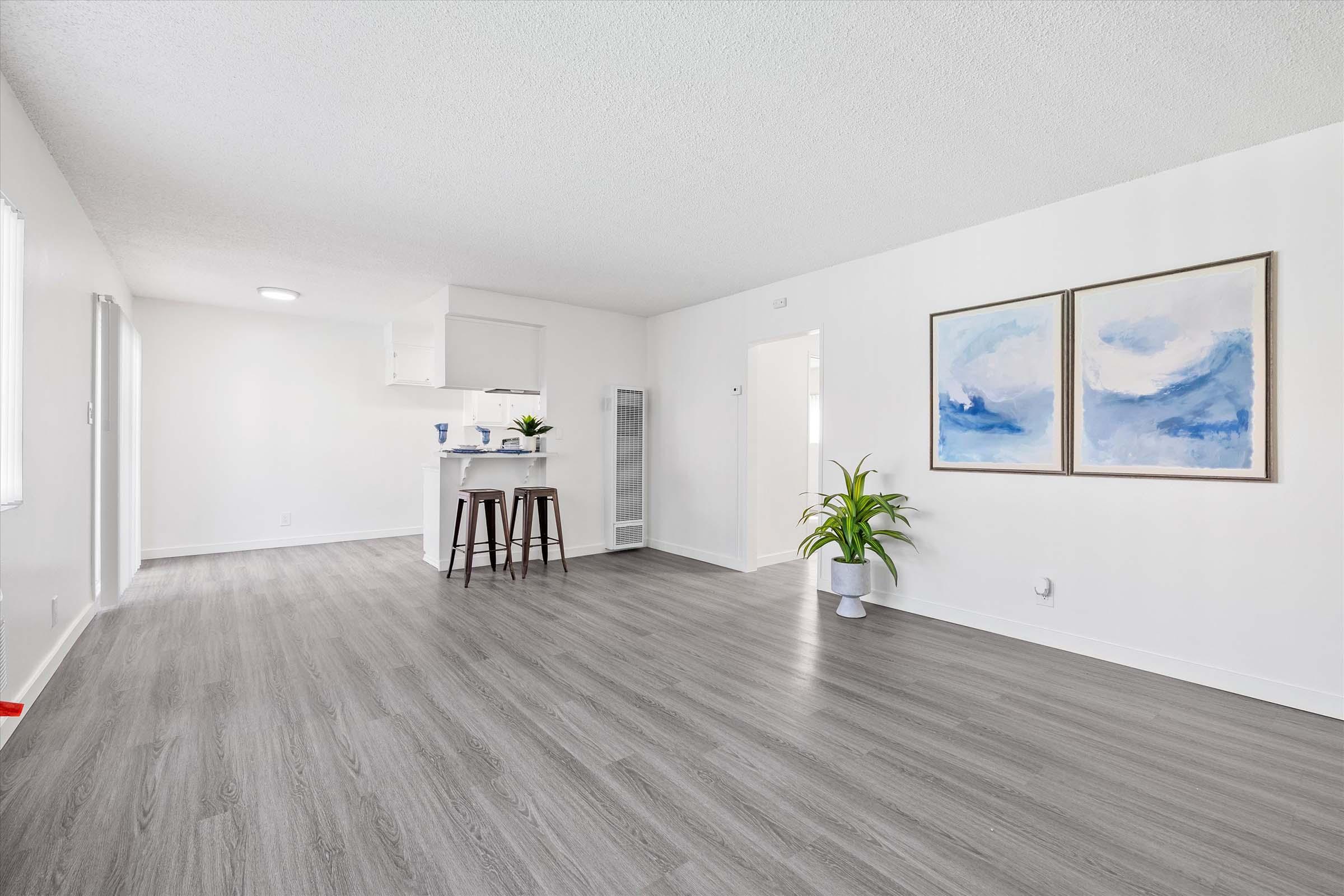 A bright, modern interior of a living space featuring light gray laminate flooring. There are two abstract paintings on the white wall, a small kitchen area with bar stools in the background, and a plant in the corner. The overall color scheme is neutral, creating a spacious and inviting atmosphere.