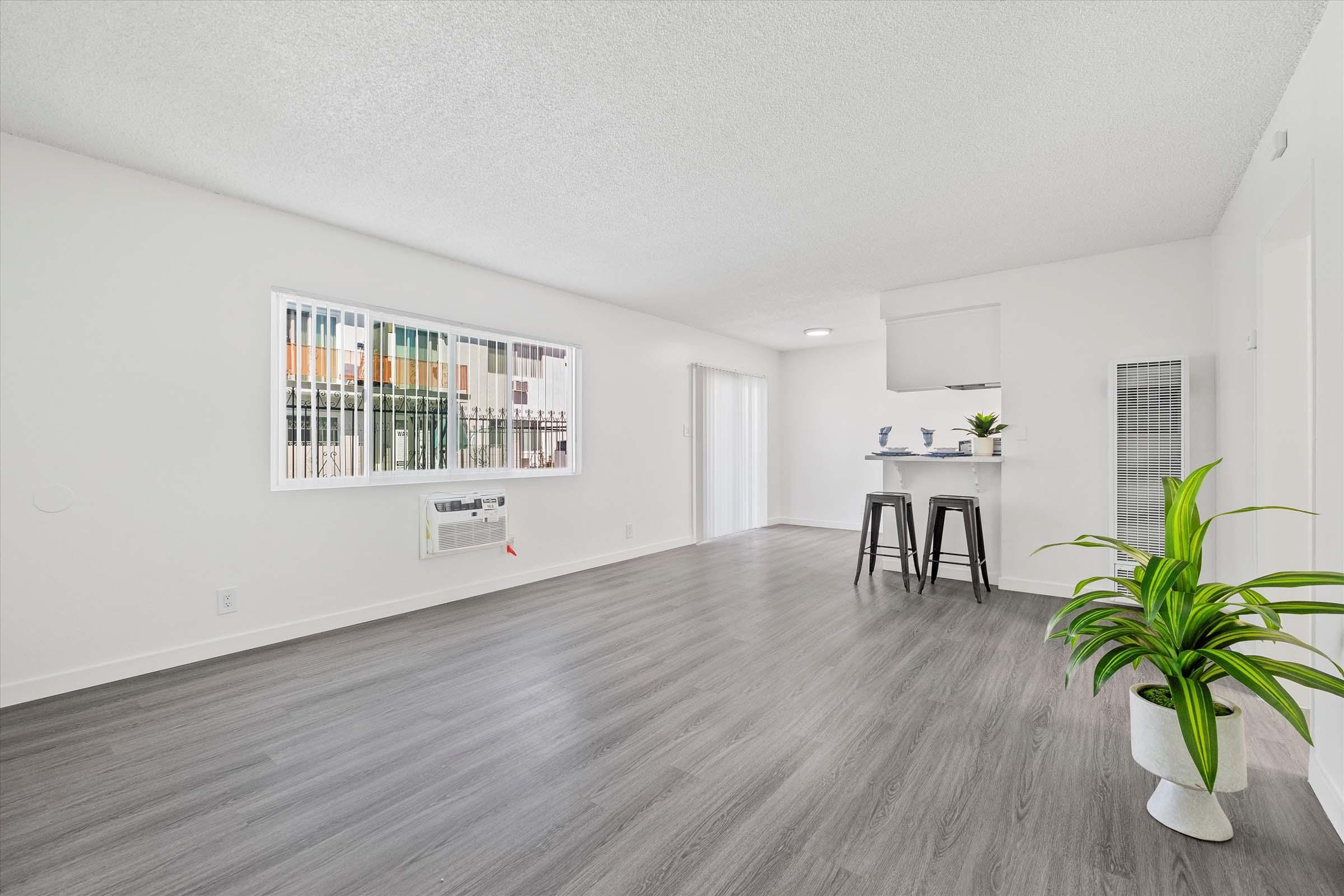 A bright, modern living room featuring gray laminate flooring, white walls, and large windows. A small bar area with two stools is visible, along with a potted plant. The room is minimally furnished, creating an open and airy atmosphere, with natural light streaming in.