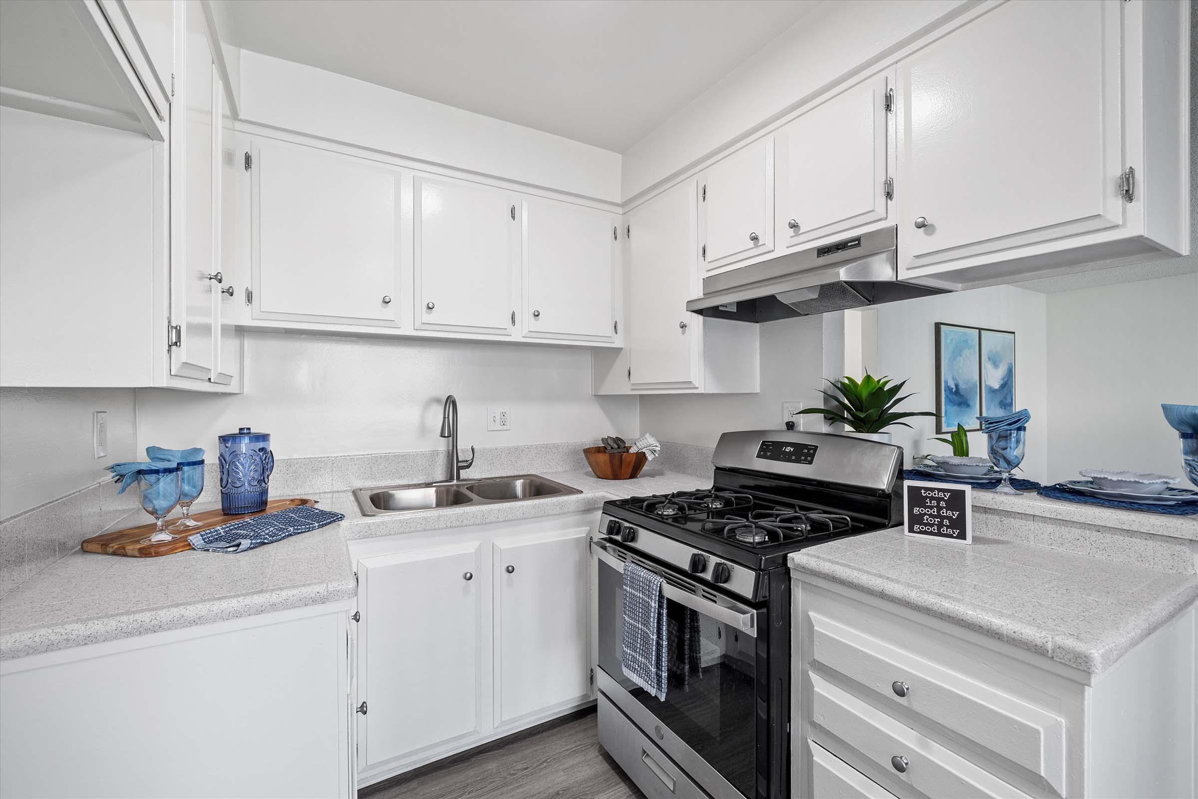 A modern kitchen featuring white cabinetry, a stainless steel gas range, and a double sink. The countertop is light-colored, complemented by a cutting board and decorative bowls. There are blue kitchen accessories, including a vase and placemat, adding a pop of color to the space.