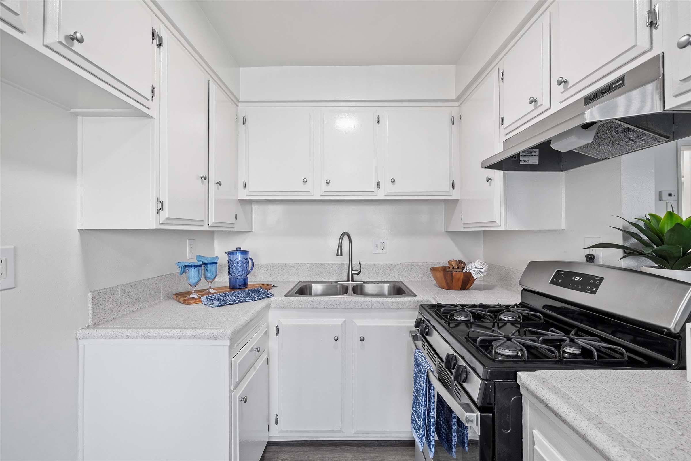 A modern kitchen featuring white cabinets, a stainless steel gas stove, and a double sink. The countertop is light gray with a bowl of fruit and blue dishware displayed. A small plant adds a touch of greenery, and there are blue kitchen towels hanging from the stove. The overall design is bright and clean.
