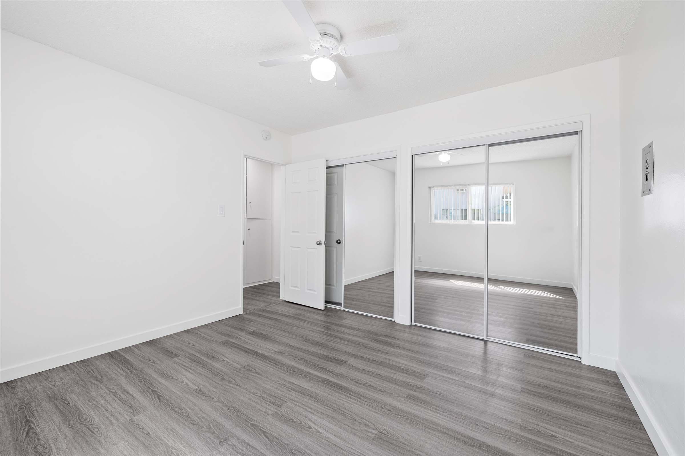 A bright, empty room with light-colored walls and sleek laminate flooring. It features a ceiling fan, a closet with sliding mirrored doors, and a window with vertical blinds allowing natural light. The space has a modern and minimalist feel, perfect for personal customization.
