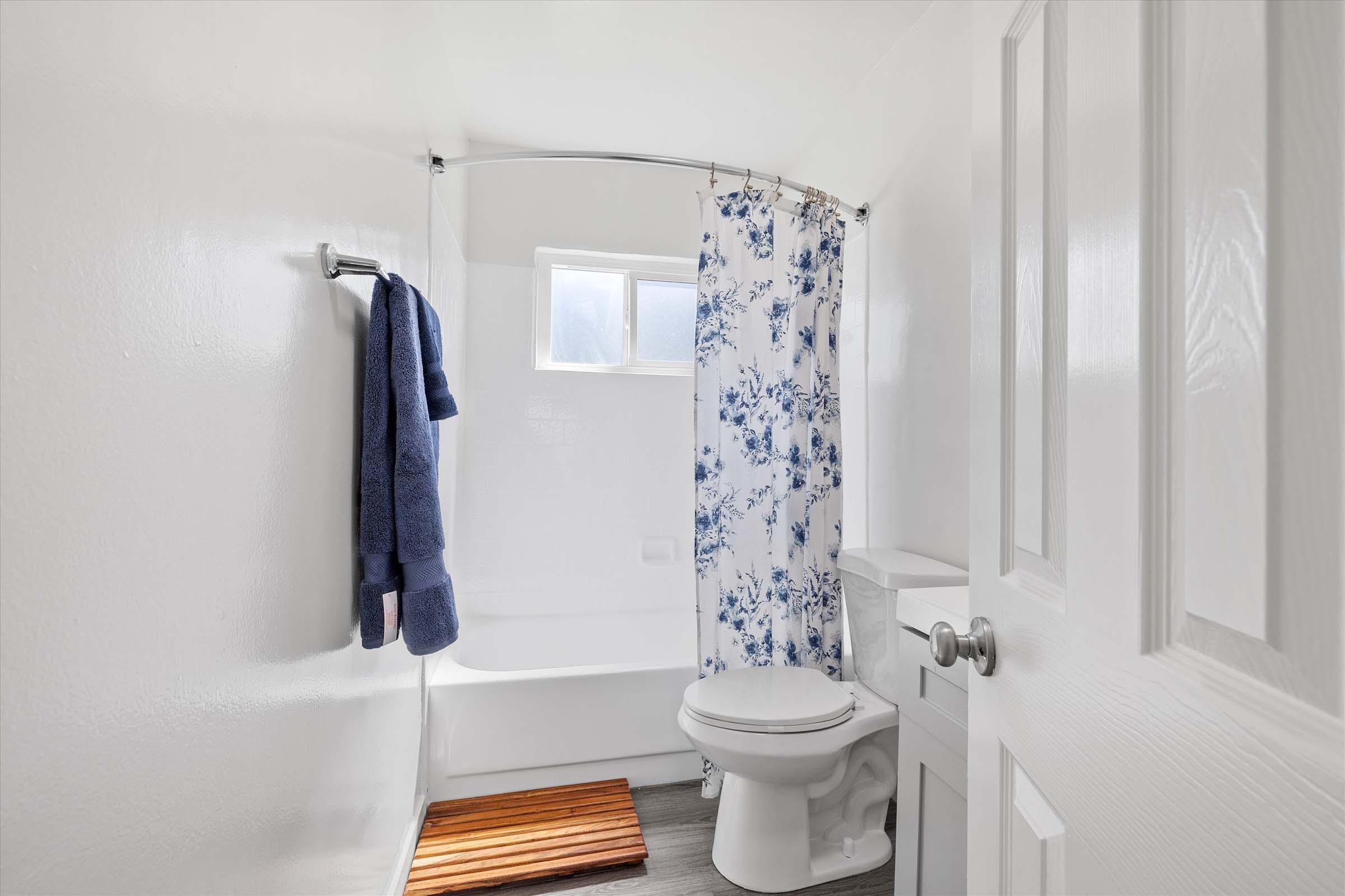 A clean, bright bathroom featuring a white bathtub with a floral shower curtain, a toilet, and a wooden bath mat. The room has a towel hanging on a rack and a small window, providing natural light. The walls are painted white, creating a fresh and airy atmosphere.