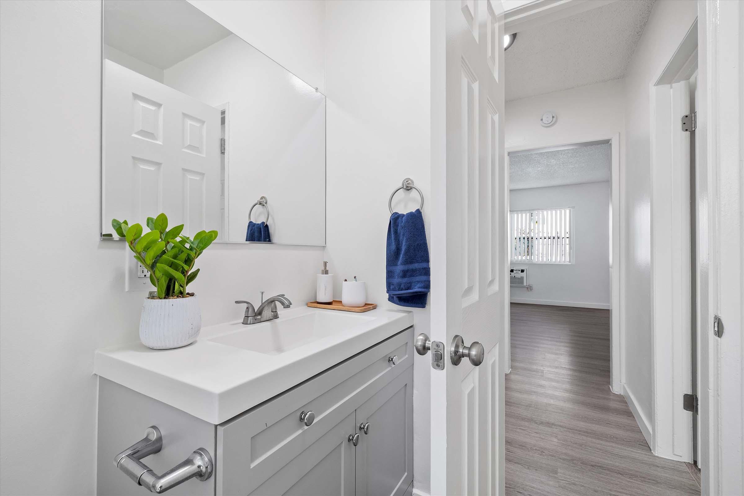 A modern bathroom featuring a white vanity with a sink, a large mirror, and a small potted plant. A blue hand towel hangs on a rack, and light-colored walls and flooring create a bright atmosphere. A door leads to another room with windows that allow natural light.