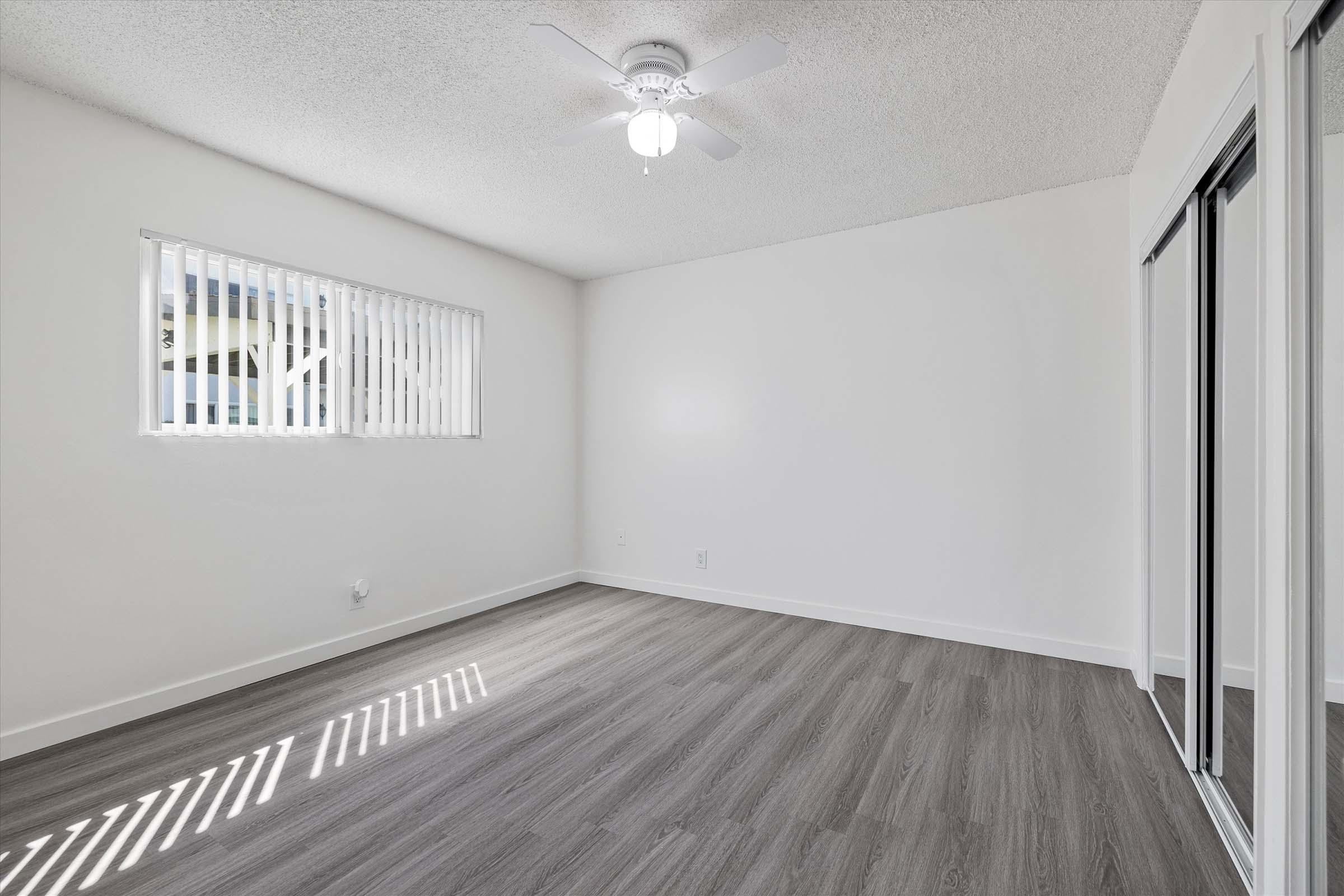 A bright, empty room with light-colored walls and a ceiling fan. It features a window with blinds, allowing natural light to enter, and has wooden-style flooring. Mirrored sliding doors are visible on one wall, and the space is clean and well-lit, creating a fresh atmosphere.
