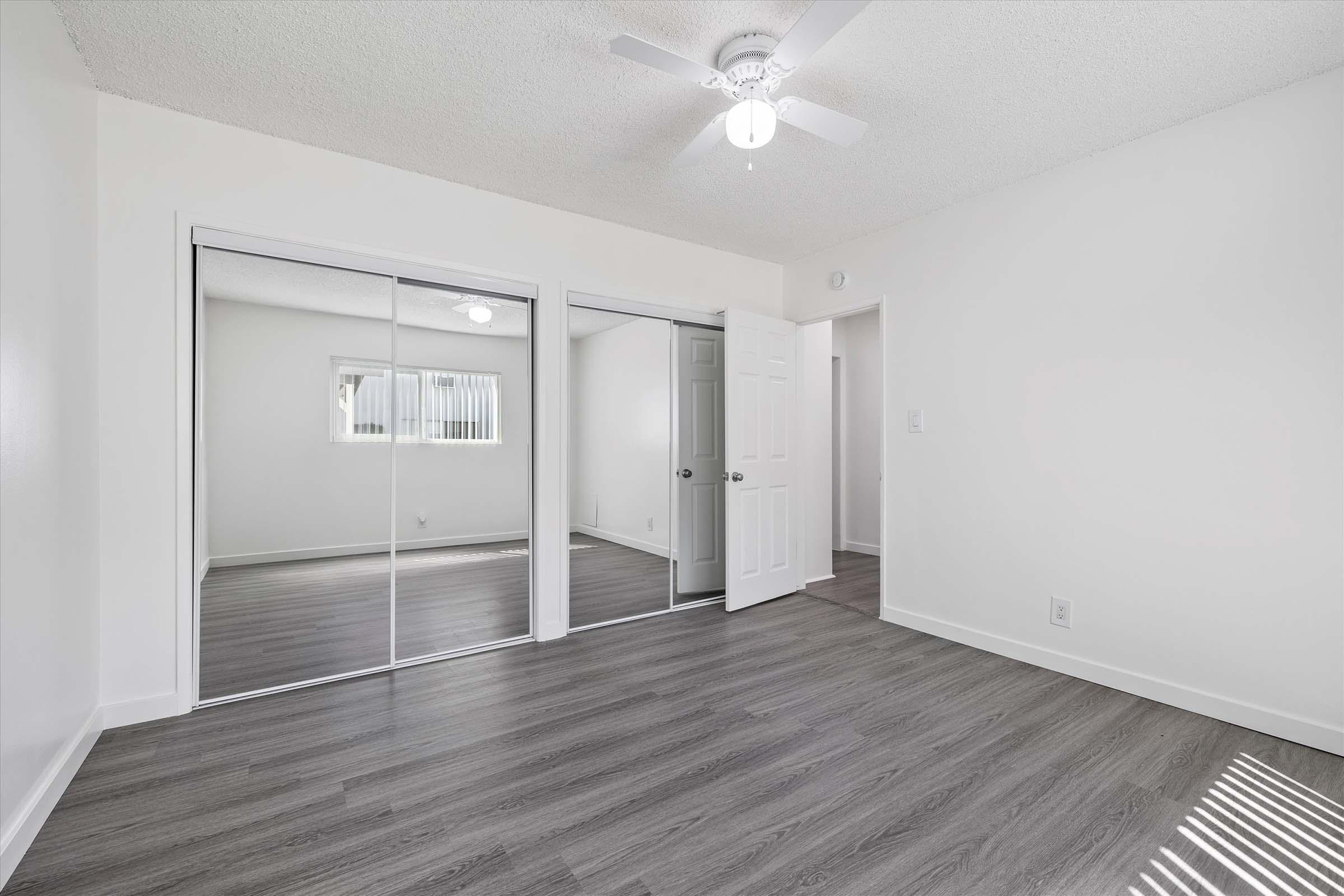 A bright and airy room featuring light gray walls and a mirrored closet. The floor is covered in sleek wood-like laminate. A ceiling fan provides airflow, and a door leads to another space. Large windows let in natural light, creating an inviting atmosphere.