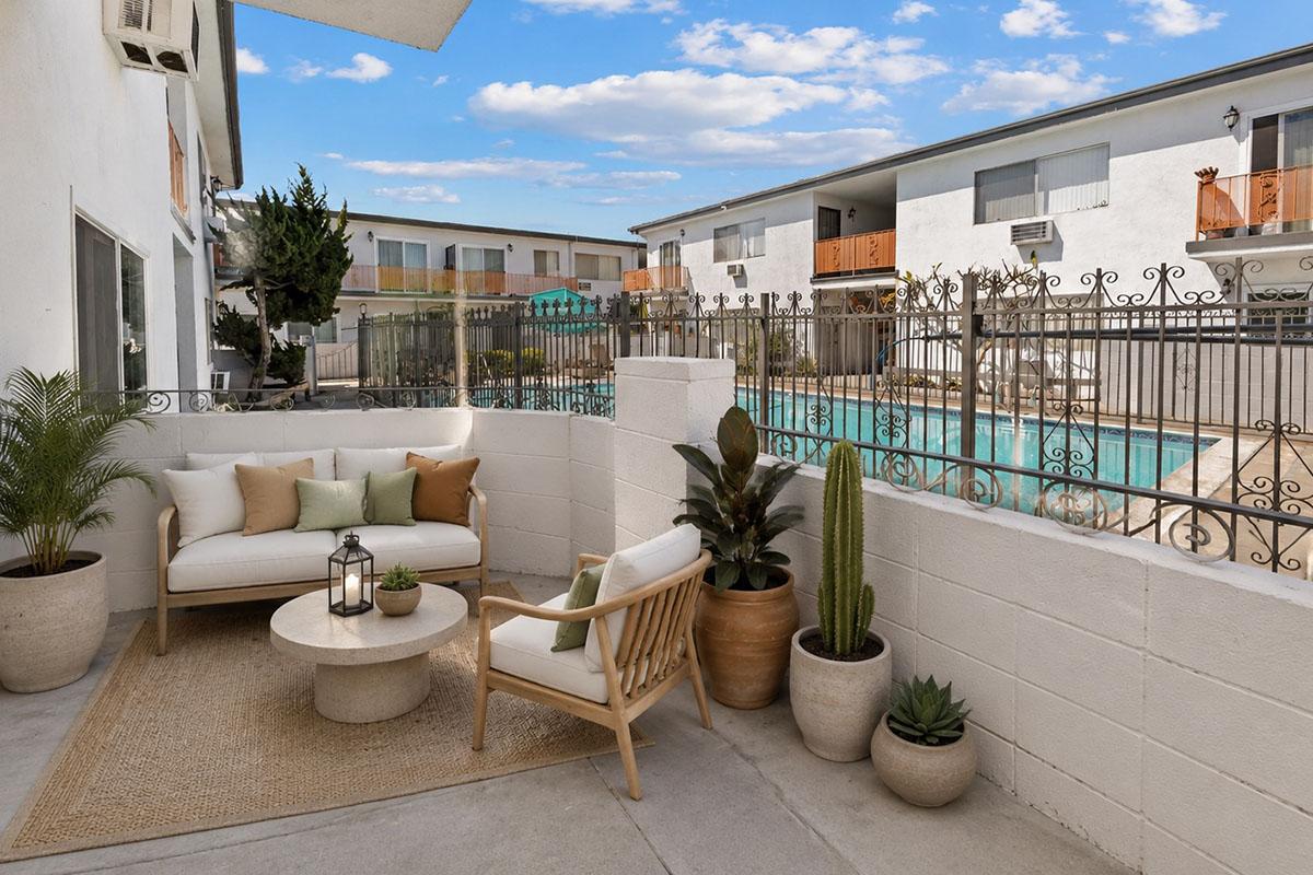 A cozy outdoor space featuring a comfortable sofa with cushions, a wooden armchair, and potted plants. The area is bordered by a decorative fence overlooking a swimming pool. The backdrop includes low-rise residential buildings and a clear blue sky, creating a serene atmosphere.