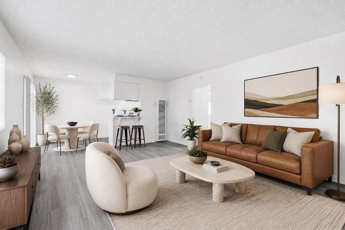 A modern living room featuring a brown leather sofa with decorative cushions, a round light-colored coffee table, and a beige rug. Nearby, there is a stylish armchair and a wooden sideboard. A dining area with a small table and chairs is visible in the background. The walls are white, and natural light fills the space.