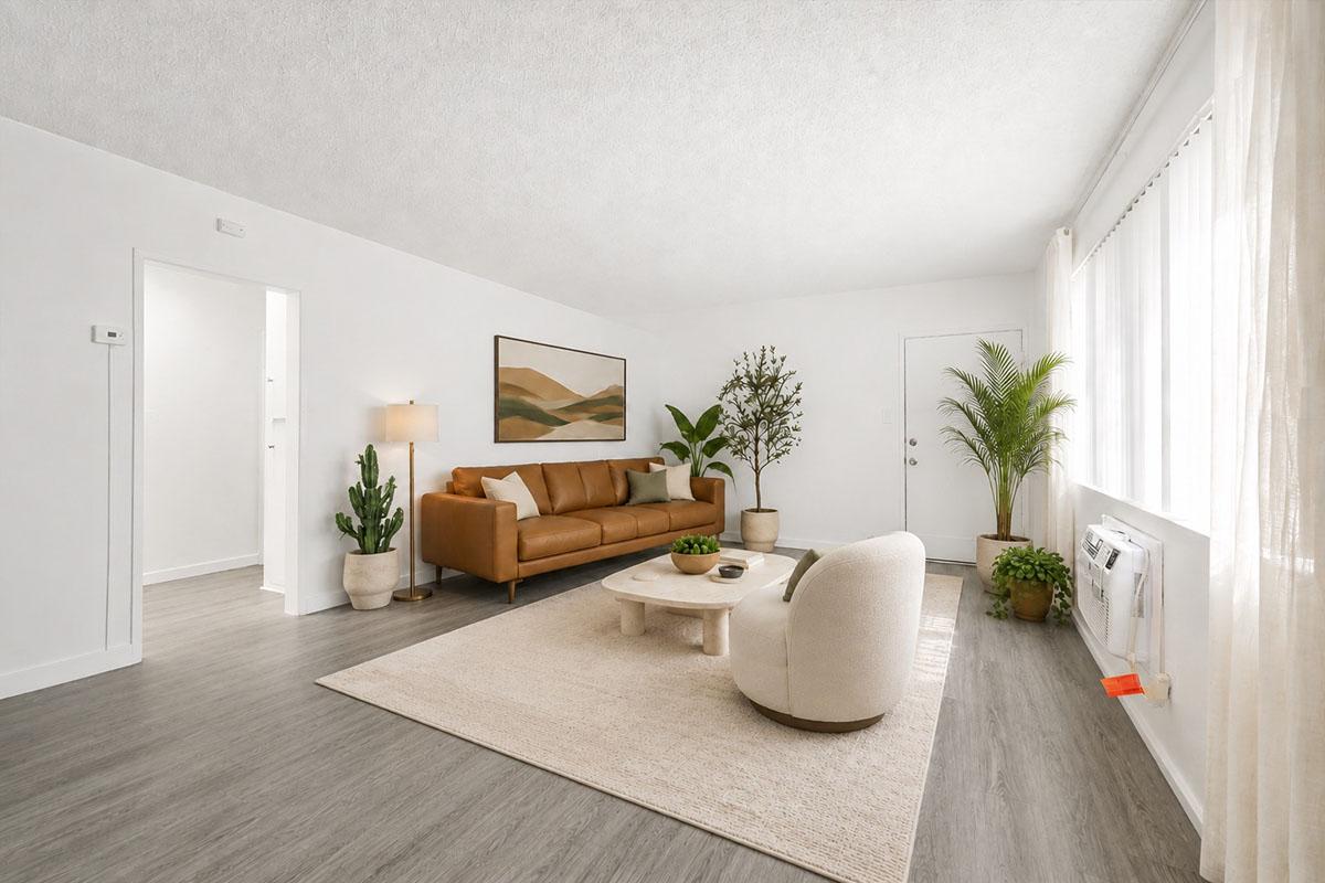 A spacious living room featuring a brown leather sofa, a light-colored coffee table, and a neutral area rug. There are potted plants and a floor lamp, with large windows allowing natural light to fill the space. The walls are painted white, and there is a modern art piece above the sofa.