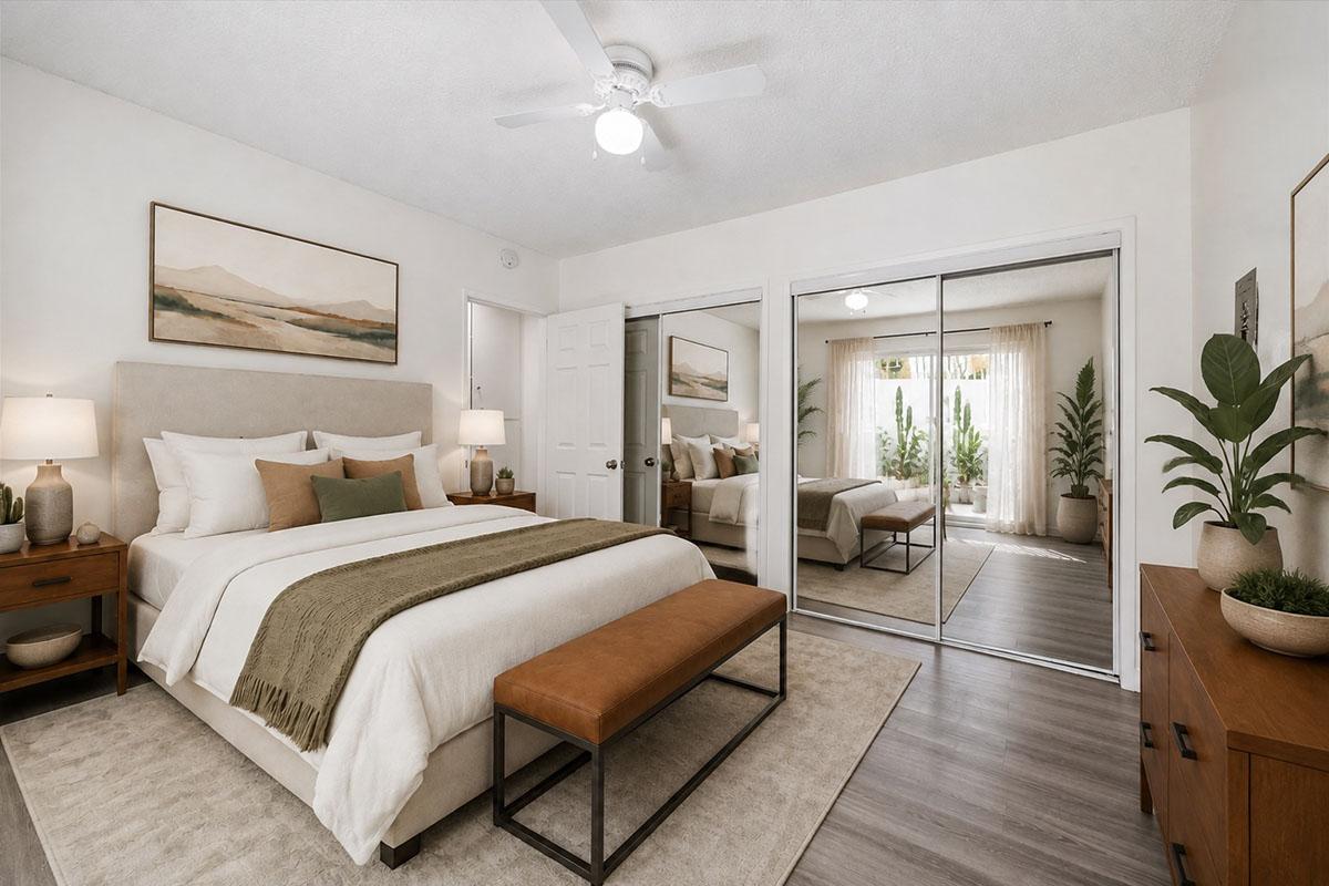 A cozy bedroom featuring a queen-sized bed with white and green bedding, flanked by two nightstands with lamps. A large mirror reflects the room, which includes a seating area with a bench, a stylish plant, and artwork on the walls. Natural light fills the space through glass doors leading to a patio.
