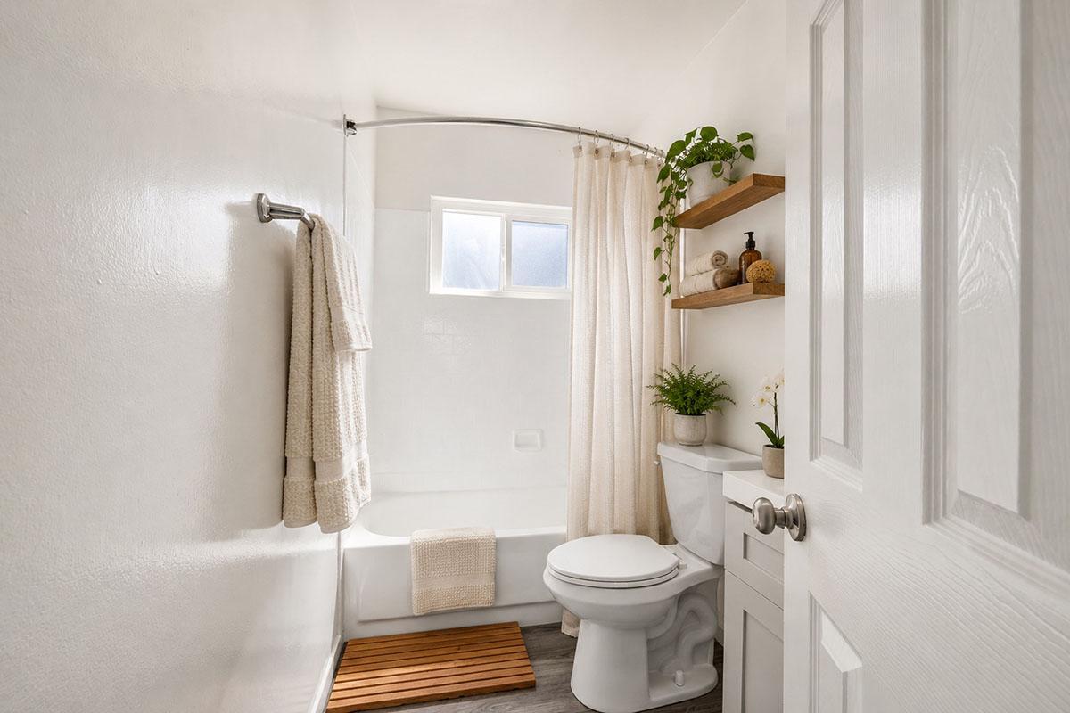 A bright, modern bathroom featuring a white tub with a shower curtain, a small window, and wooden shelves with decorative items. There is a potted plant on the shelf and a towel hanging on the wall. The floor is covered with a wooden mat, creating a clean and inviting space.