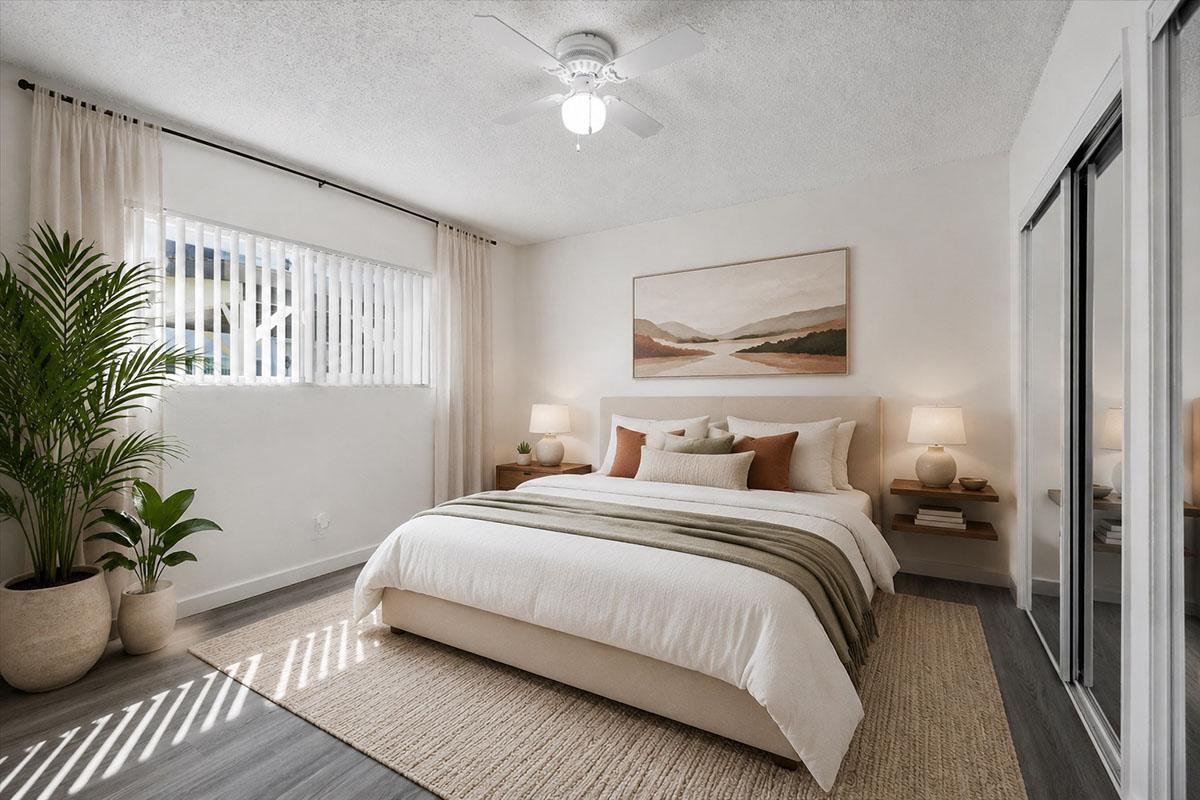 A bright and airy bedroom featuring a king-size bed with a light-colored duvet and accent pillows. There are two bedside tables with lamps, a large painting above the bed, and a potted plant in the corner. Natural light streams in through a window with horizontal blinds, illuminating the neutral-toned decor.