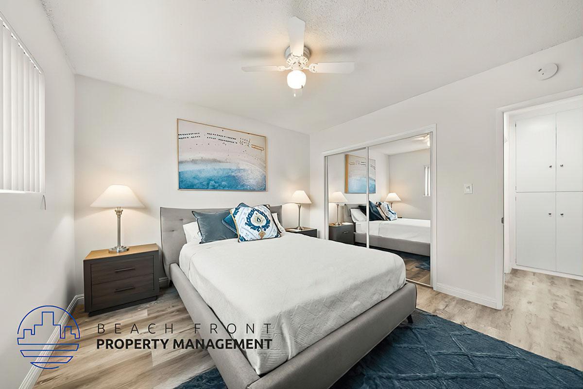A bright and modern bedroom featuring a queen-sized bed with a decorative blue and white pillow. There are two bedside lamps on wooden nightstands, a large mirror reflecting the room, and a ceiling fan. The walls are light-colored, and an abstract painting hangs above the bed, adding a touch of decor.