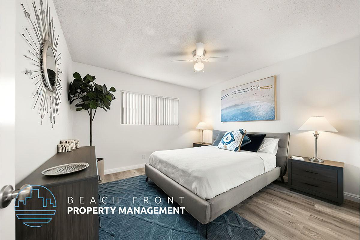 A bright and modern bedroom featuring a queen-sized bed with blue and white bedding, a decorative wall art piece depicting ocean waves, a nightstand with a lamp on each side, a small plant in the corner, and a light wood floor. The room has natural light filtering through a window with blinds.