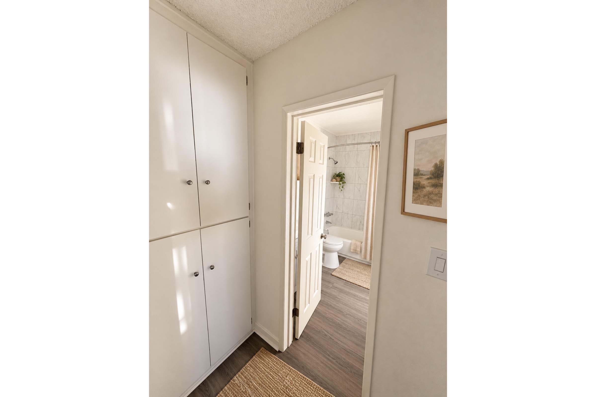 A view of a bathroom doorway from a hallway. The hallway features white cabinets with knobs. The bathroom visible through the doorway has light-colored walls, a toilet, and a shower curtain. A framed picture is on the wall near the door, and the floor is covered with a woven mat.