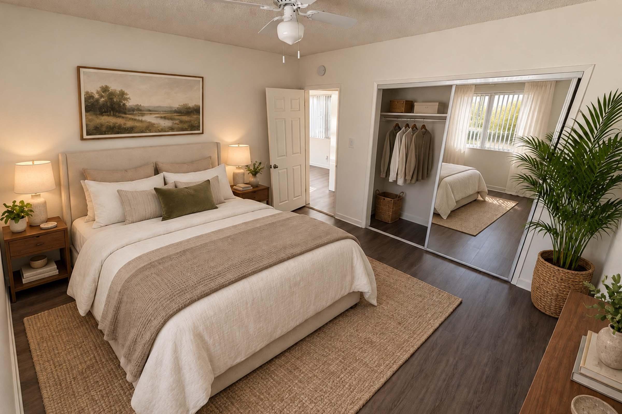 A cozy, well-lit bedroom featuring a neatly made bed with neutral-toned bedding. There are bedside tables with lamps on either side, potted plants, and a large mirror reflecting a closet with hanging clothes. The walls are painted a soft color, contributing to a warm and inviting atmosphere.