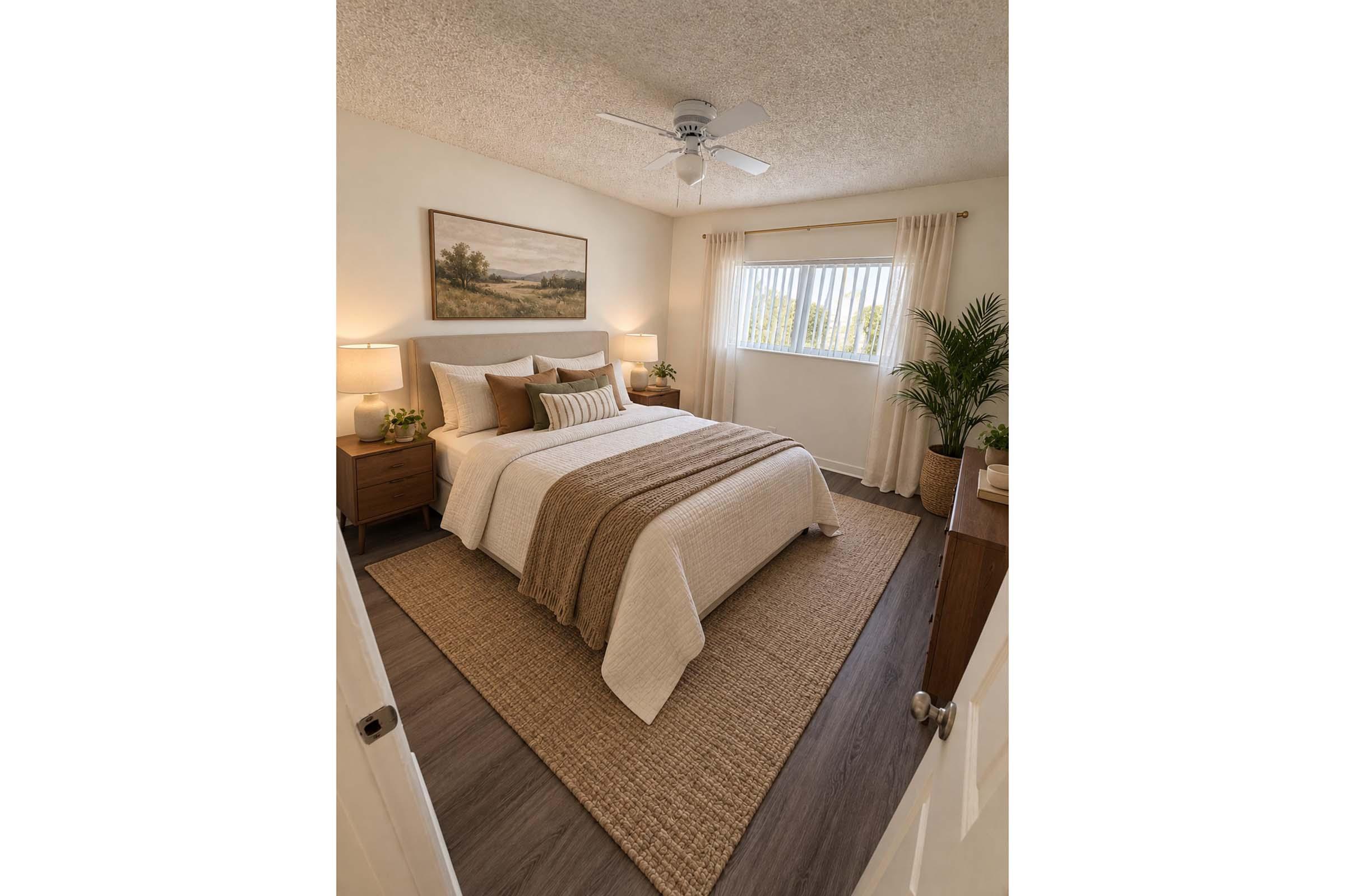 A cozy bedroom with a queen bed dressed in a light-colored quilt and decorative pillows. There are two bedside lamps on wooden nightstands, a large window with cream curtains, and a potted plant. A tapestry of a landscape hangs above the bed, and a woven rug covers the floor. The walls are painted a soft white, creating a warm ambiance.