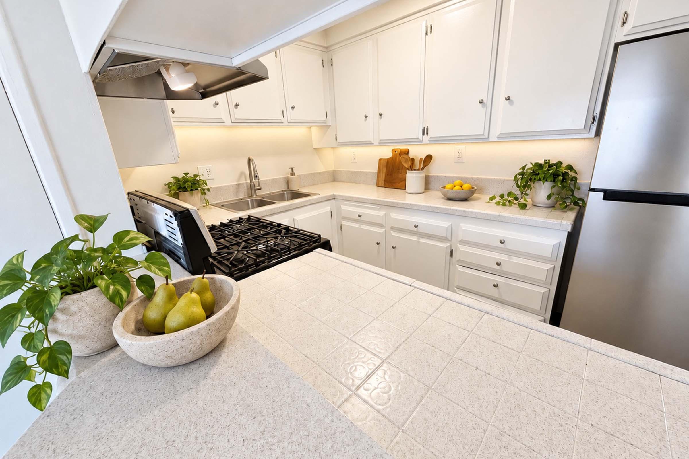 A bright and modern kitchen featuring white cabinets and a gray countertop. A gas stove is visible, along with a stainless steel fridge. A bowl of green pears sits on the countertop, accompanied by potted plants. Sunlight illuminates the space, enhancing the warm and inviting atmosphere.