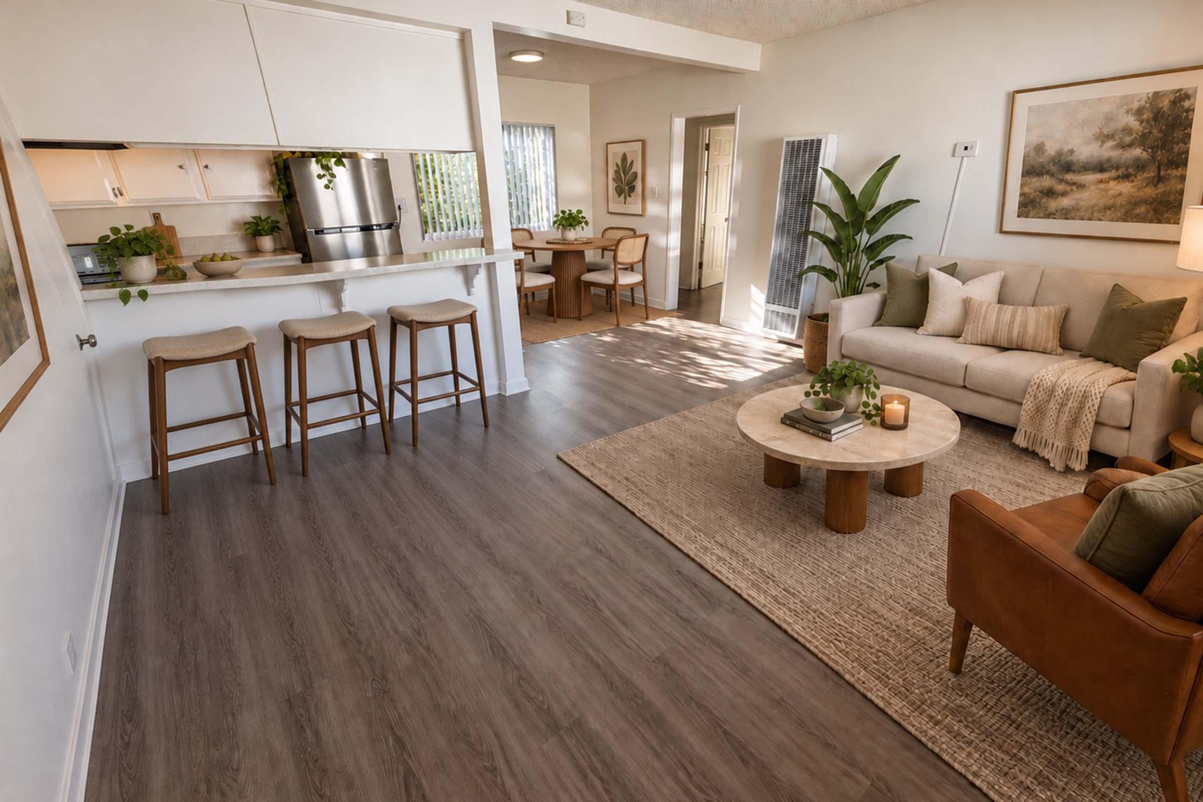 A spacious and modern living area featuring a cozy sofa with decorative pillows, a round coffee table, and a rug. A kitchen bar with three stools is visible, along with indoor plants and artwork on the walls. Natural light fills the room, creating a warm and inviting ambiance.
