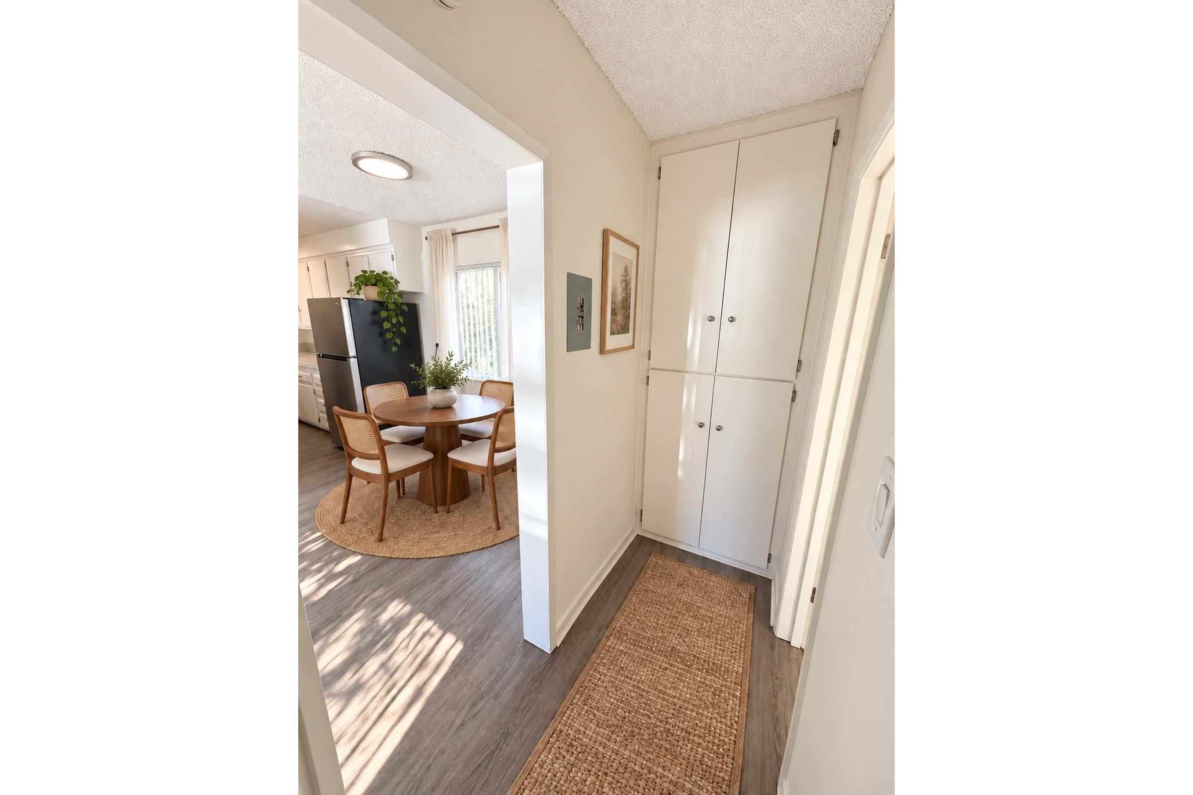A well-lit hallway leads to a kitchen area, featuring a round dining table with wooden chairs and a large rug. On the left, there is a tall white storage cabinet, and light-colored walls enhance the airy feel. A small entrance mat is positioned at the hallway's entrance.