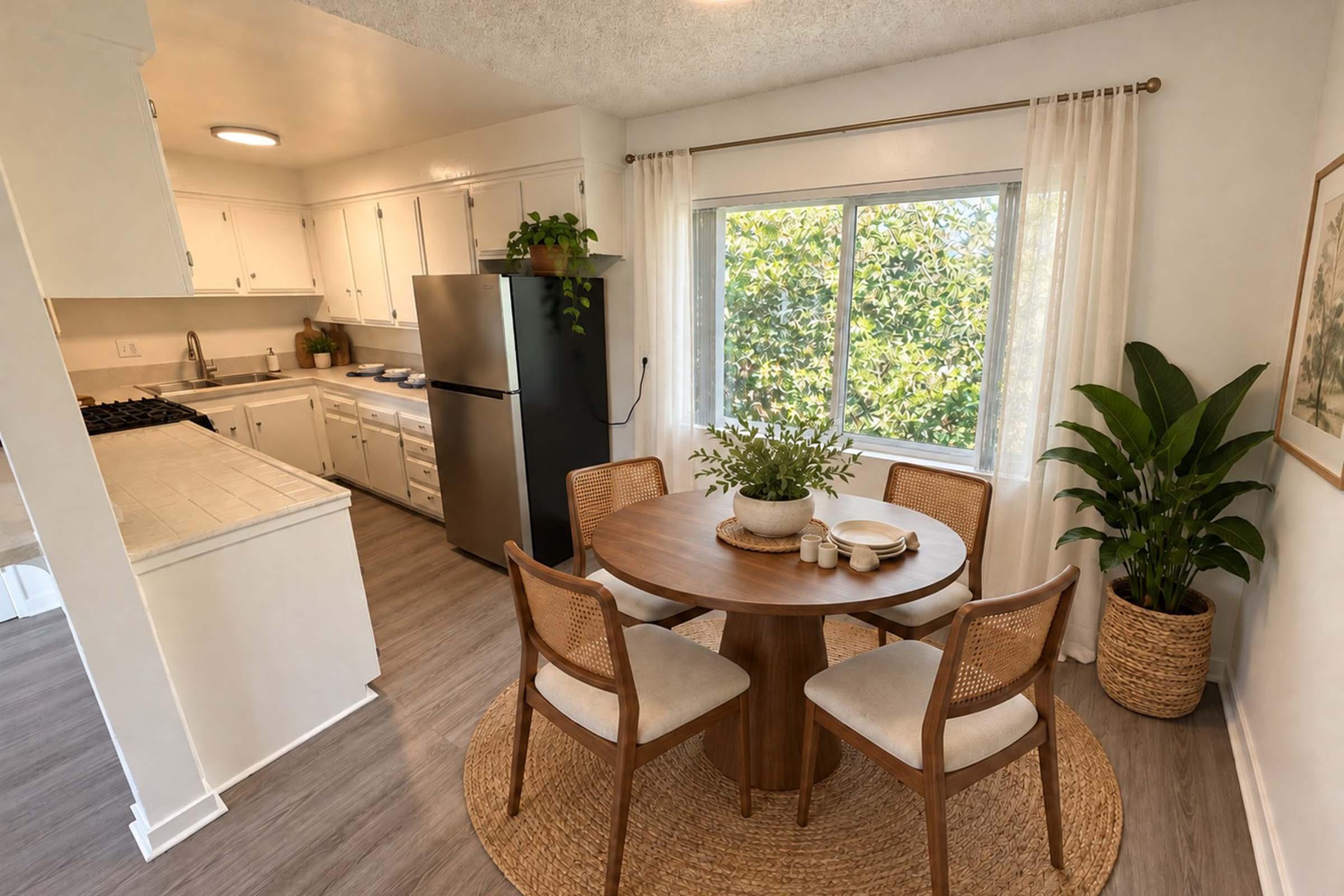 A bright, inviting kitchen and dining area featuring a round wooden table with four chairs, a potted plant, and a decorative bowl. The kitchen appliances, including a black refrigerator, are visible, along with white cabinets and a view of greenery through a large window. Natural light enhances the cozy atmosphere.