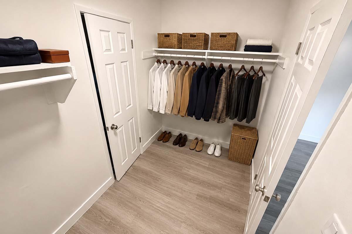 A well-organized closet featuring a variety of clothing items hung on a rack, including several shirts and a suit. Below, there are neatly arranged shoes and baskets. The walls are painted white, and the floor is light wood. Two white doors are visible, adding to the clean and tidy appearance.