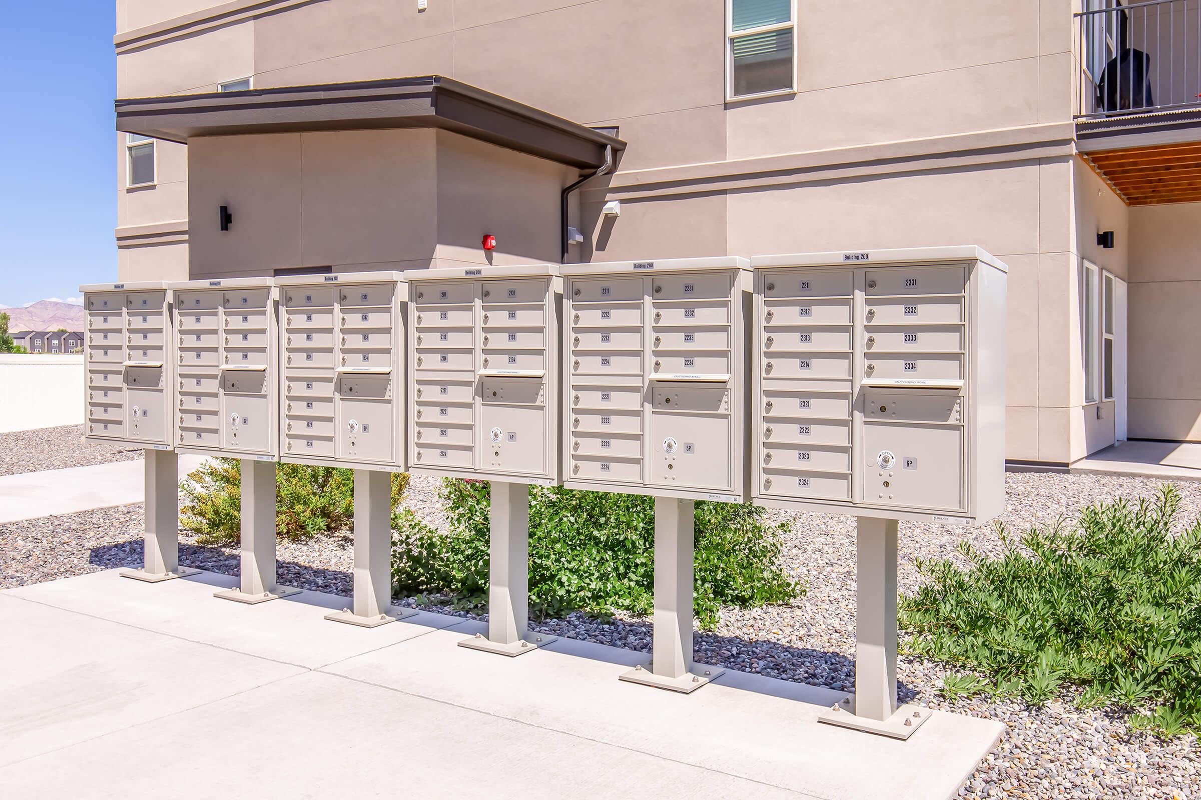 Row of metal mailboxes standing on a sidewalk outside a modern apartment building, surrounded by gravel and low greenery. The building features a light-colored exterior and a balcony above the mailboxes. Bright blue sky overhead indicates a sunny day.