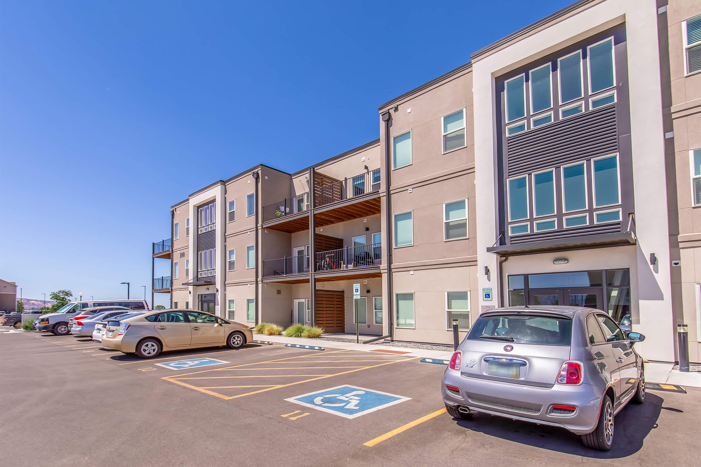 Modern apartment building with multiple units, featuring balconies and large windows. There are designated parking spaces, including a handicap spot, in the foreground. Several cars are parked, and the sky is clear and blue, indicating a bright day.