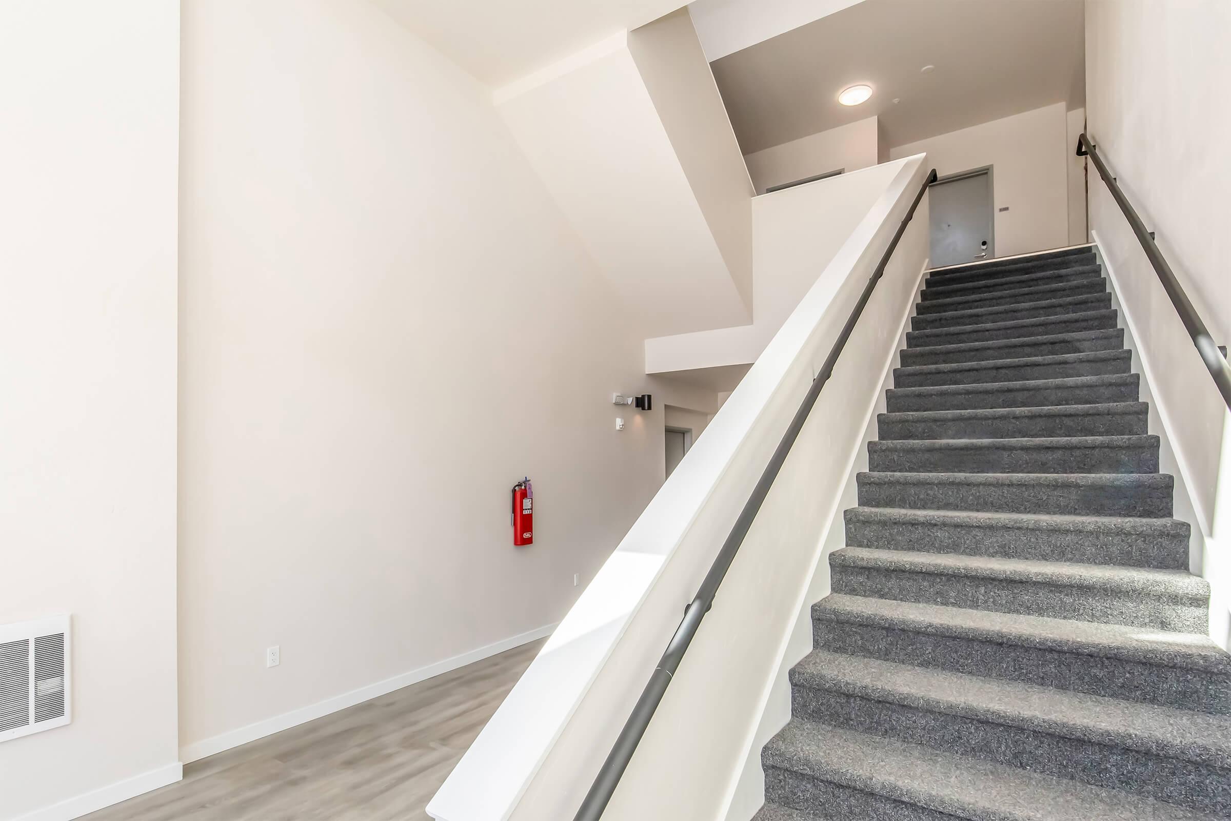 A well-lit, modern staircase with gray carpeted steps leading up. The walls are painted a light color, and there is a fire extinguisher mounted on the wall. The landing above features an open space with a door and a minimalistic design. The area feels spacious and new.