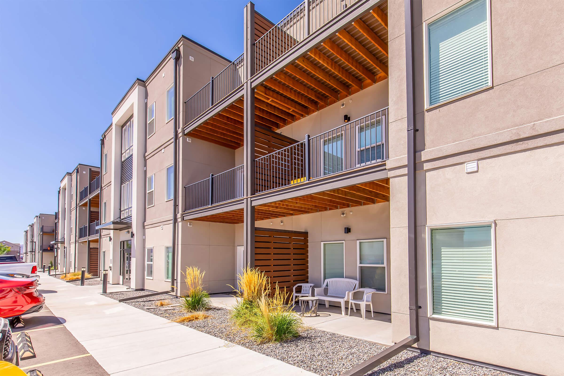A modern apartment building featuring multiple units with balconies. The facade is a mix of light-colored walls and wooden accents. There are outdoor seating areas and landscaped patches with low-maintenance plants along a pathway. Bright blue skies provide a vibrant backdrop.