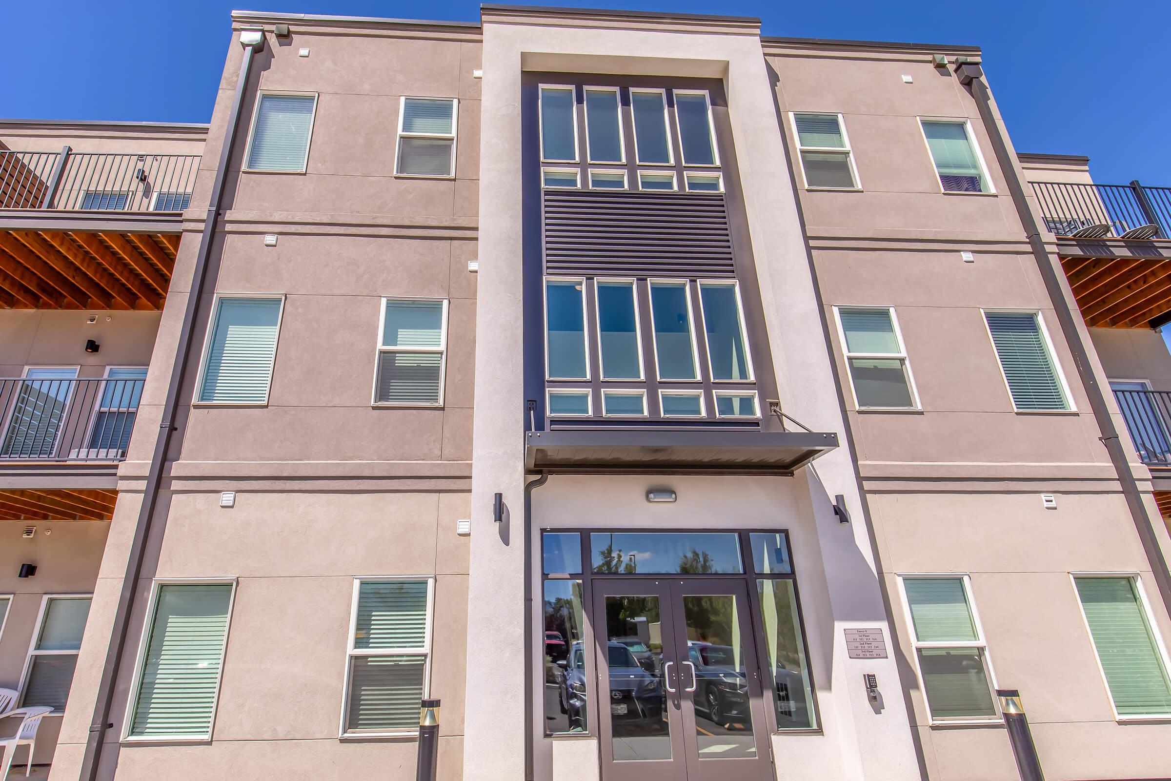 Modern multi-story apartment building with a light-colored exterior, featuring large windows and a prominent entrance. The façade includes a mix of textures and materials, with a balcony on the upper floor and a welcoming entryway. Bright blue sky complements the structure.