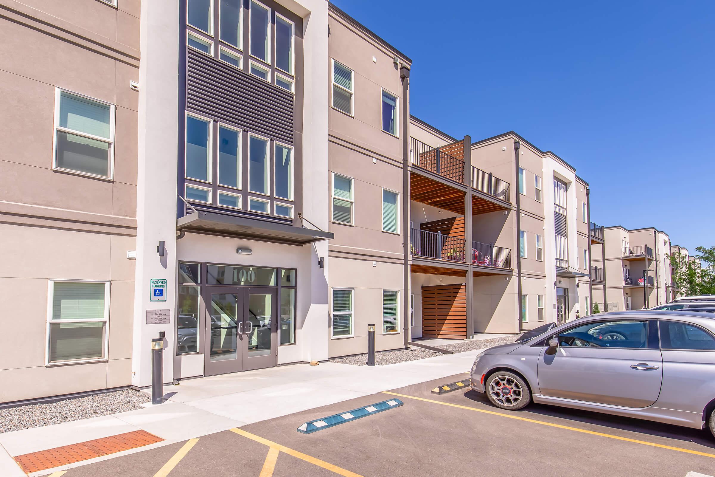 Modern apartment building with a light gray facade and large windows, featuring two stories and balconies. The entrance is centrally located with a glass door. A parking lot is visible in front, accommodating several vehicles, including a silver car. The sky is clear and blue.