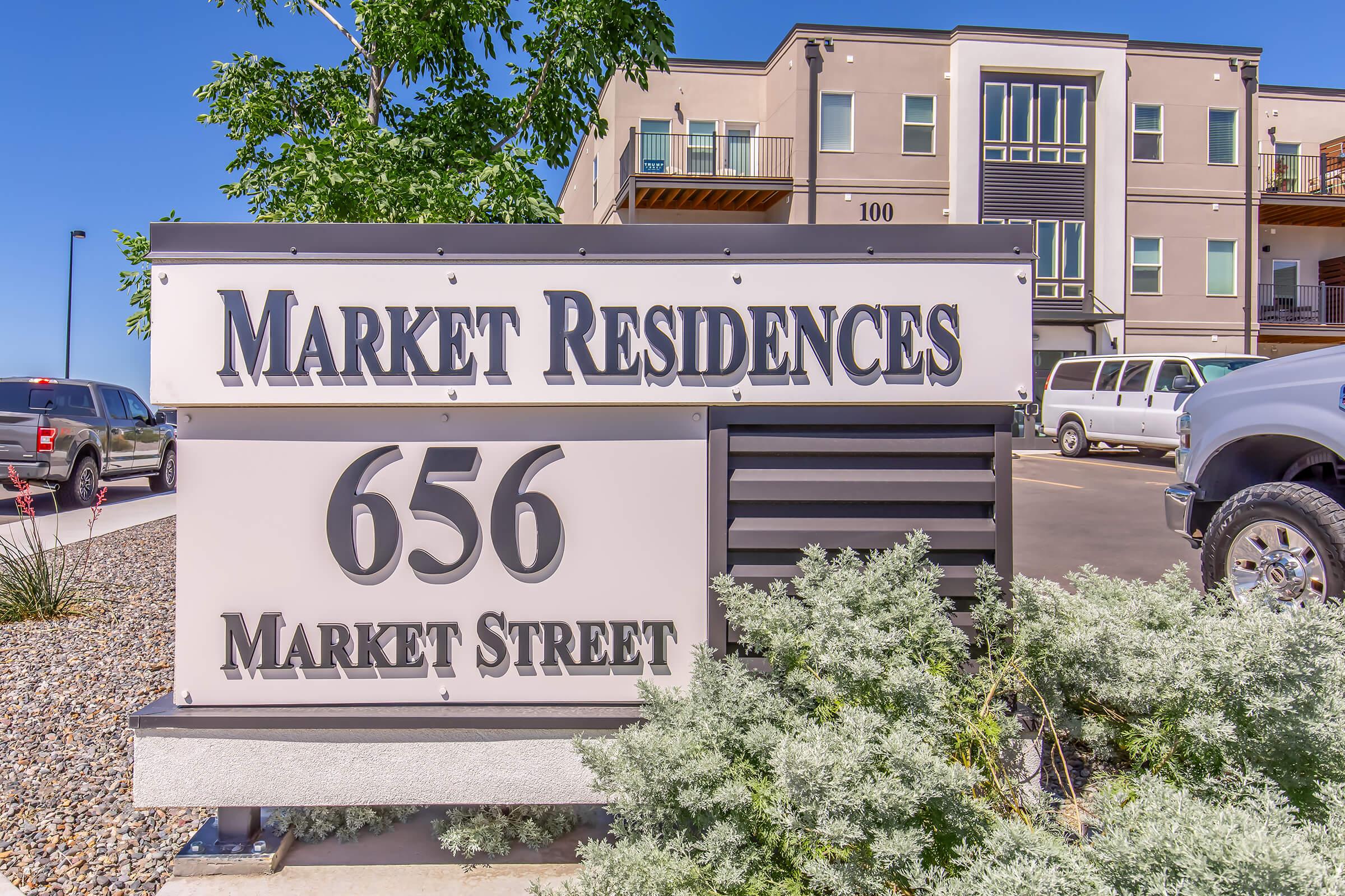 Sign for Market Residences located at 656 Market Street. The sign features large, bold lettering and is surrounded by decorative landscaping. In the background, there are modern apartment buildings, a clear blue sky, and parked vehicles.