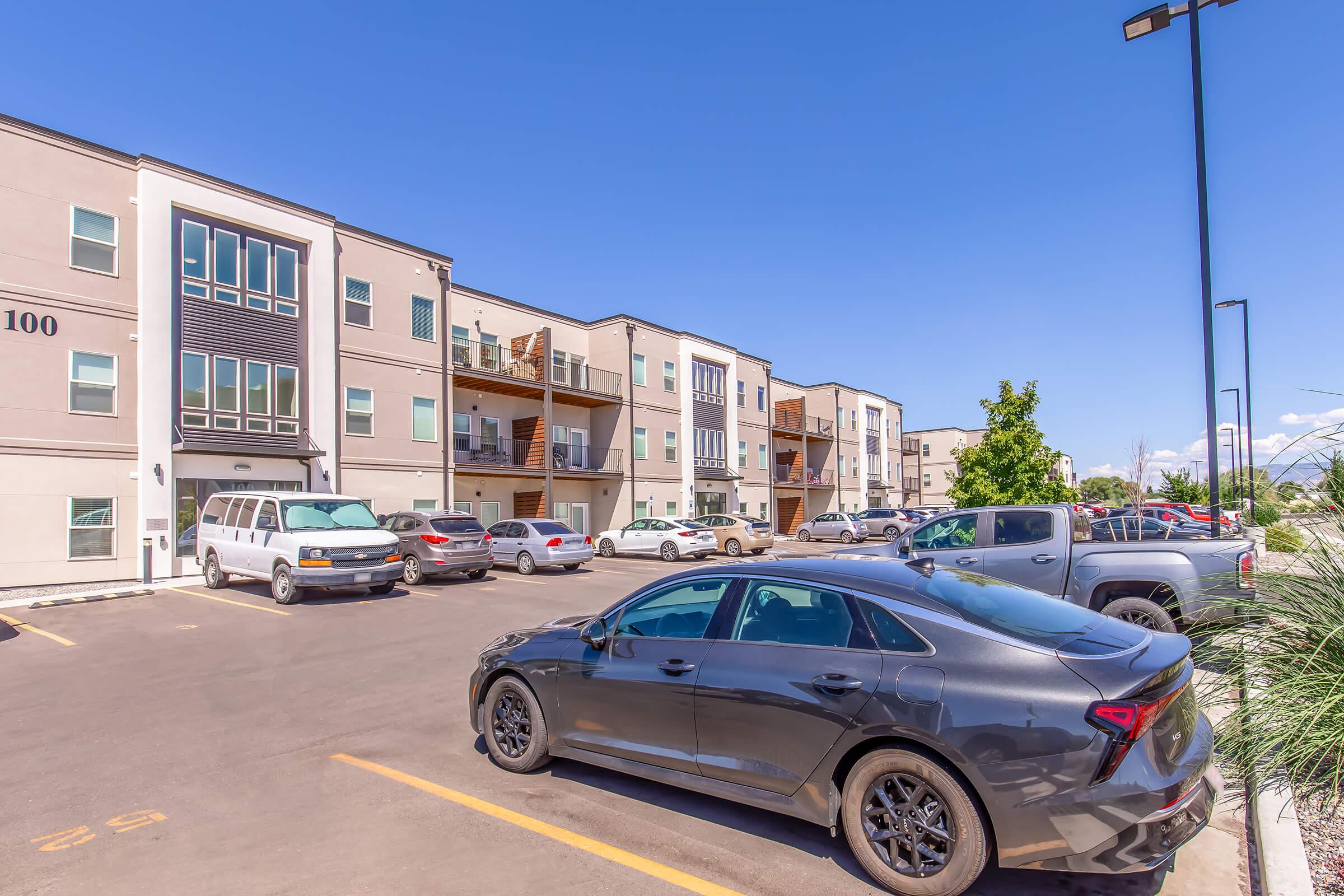 A view of a modern apartment complex featuring multiple two-story buildings with balconies, a parking lot filled with vehicles including a black car in the foreground, and a clear blue sky overhead. The scene conveys a suburban living environment with ample parking space.