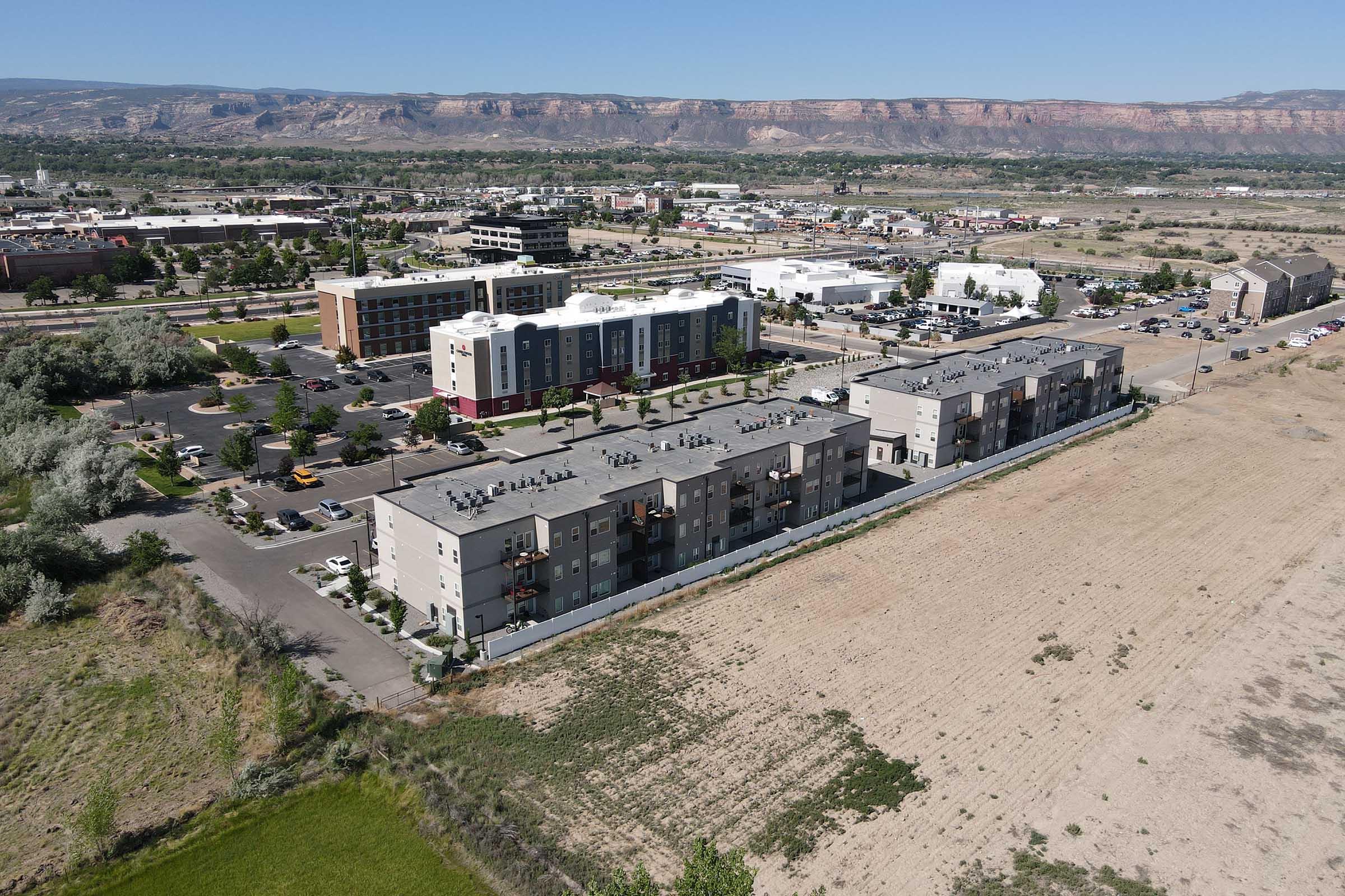 Aerial view of a suburban area featuring multiple buildings, including low-rise apartment complexes and commercial structures, set against a backdrop of mountains. The foreground shows an empty field, while the parking lots are filled with vehicles. The landscape appears sunny and clear.