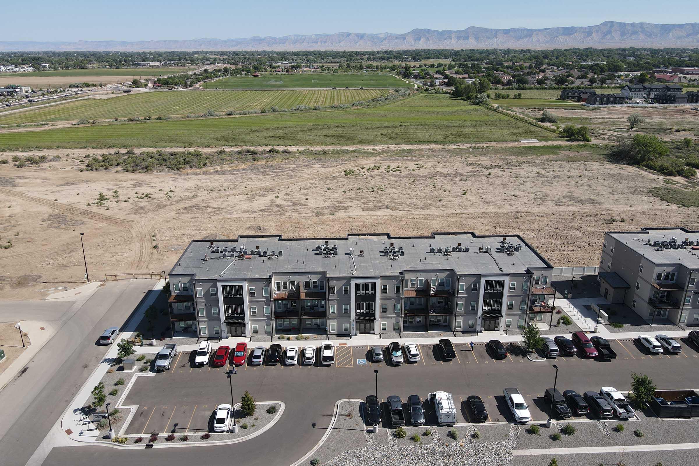 Aerial view of a residential complex with multiple two-story buildings featuring balconies and solar panels. In the foreground, there is a parking lot filled with various vehicles. In the background, fields and mountains stretch out under a clear blue sky, highlighting the rural landscape surrounding the development.