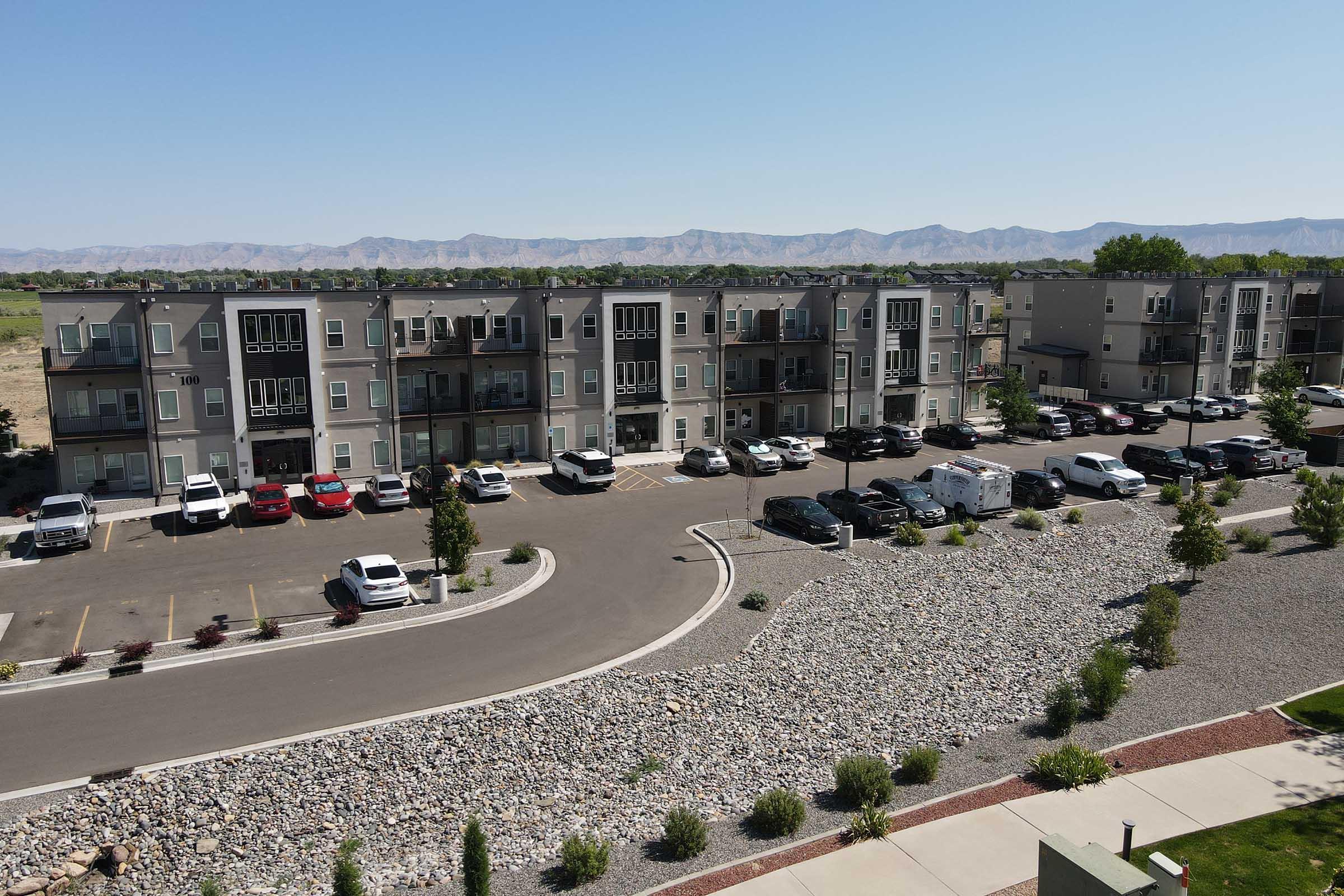 Aerial view of a residential apartment complex with multiple buildings, parking spaces filled with cars, and a landscaped area featuring gravel and greenery. In the background, mountains are visible under a clear blue sky.