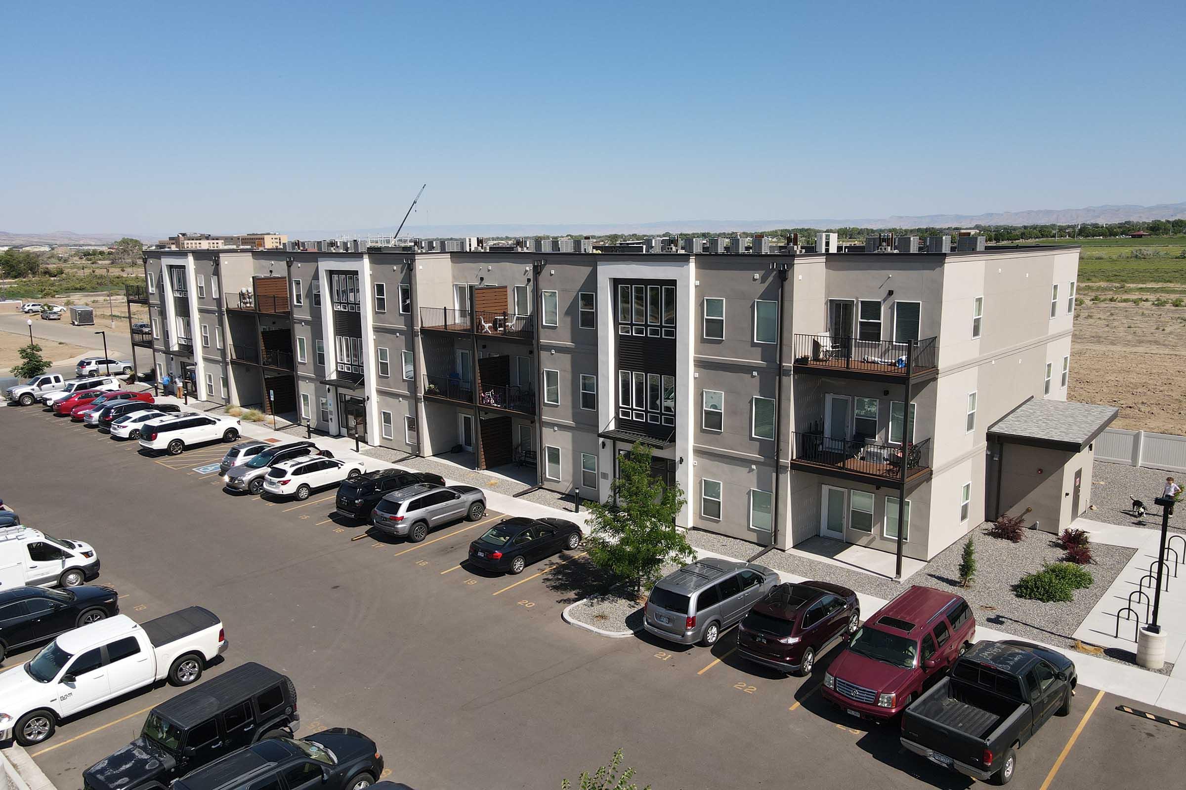 A modern multi-story apartment building with a row of parked cars in front. The building features balconies and a clean, contemporary design, surrounded by open land and distant hills under a clear blue sky.