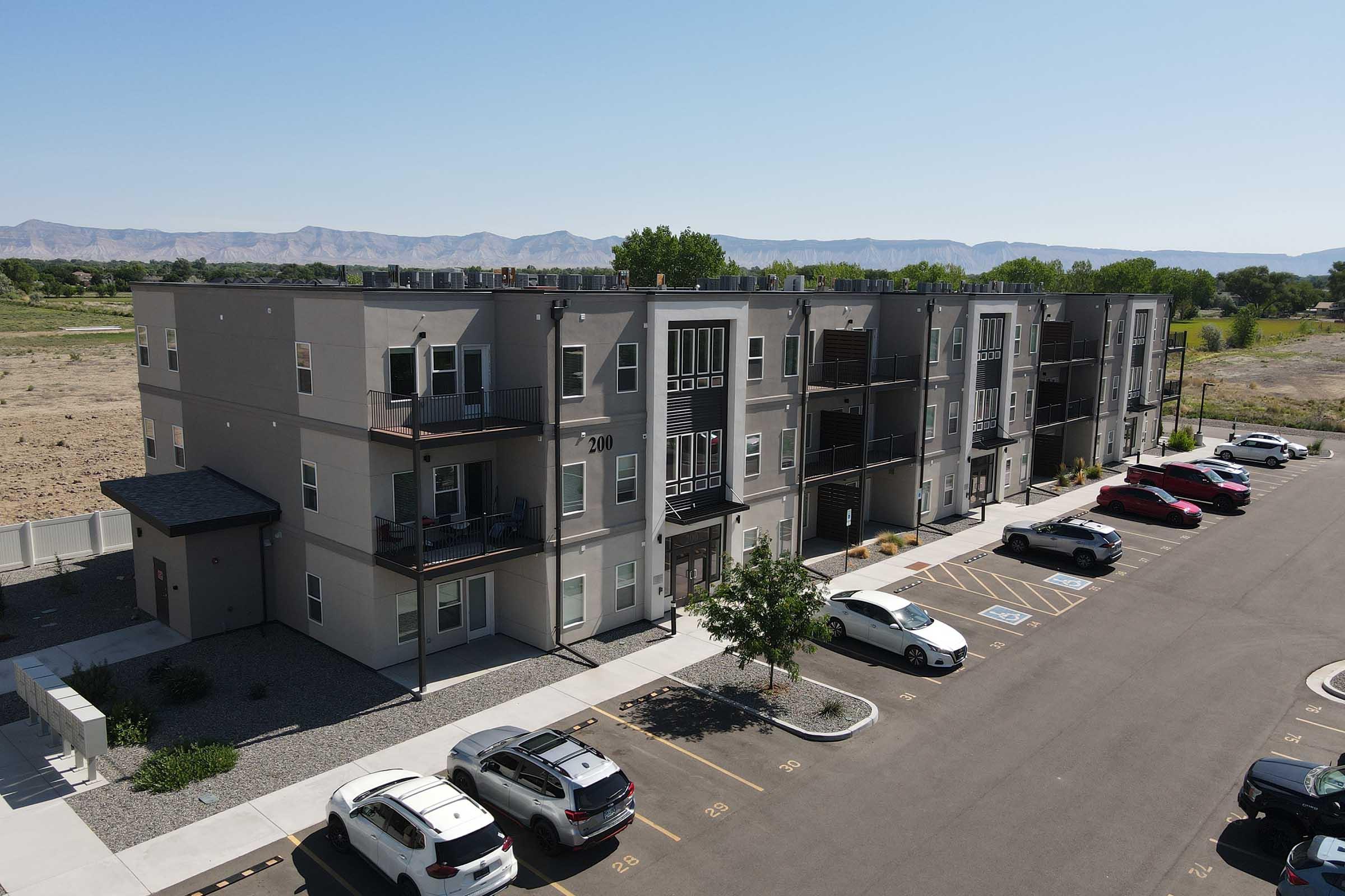 A modern, multi-story apartment building featuring gray exterior walls and several balconies. The structure is surrounded by a parking lot with multiple parked vehicles. In the background, there are open fields and mountains under a clear blue sky. The setting appears to be suburban.