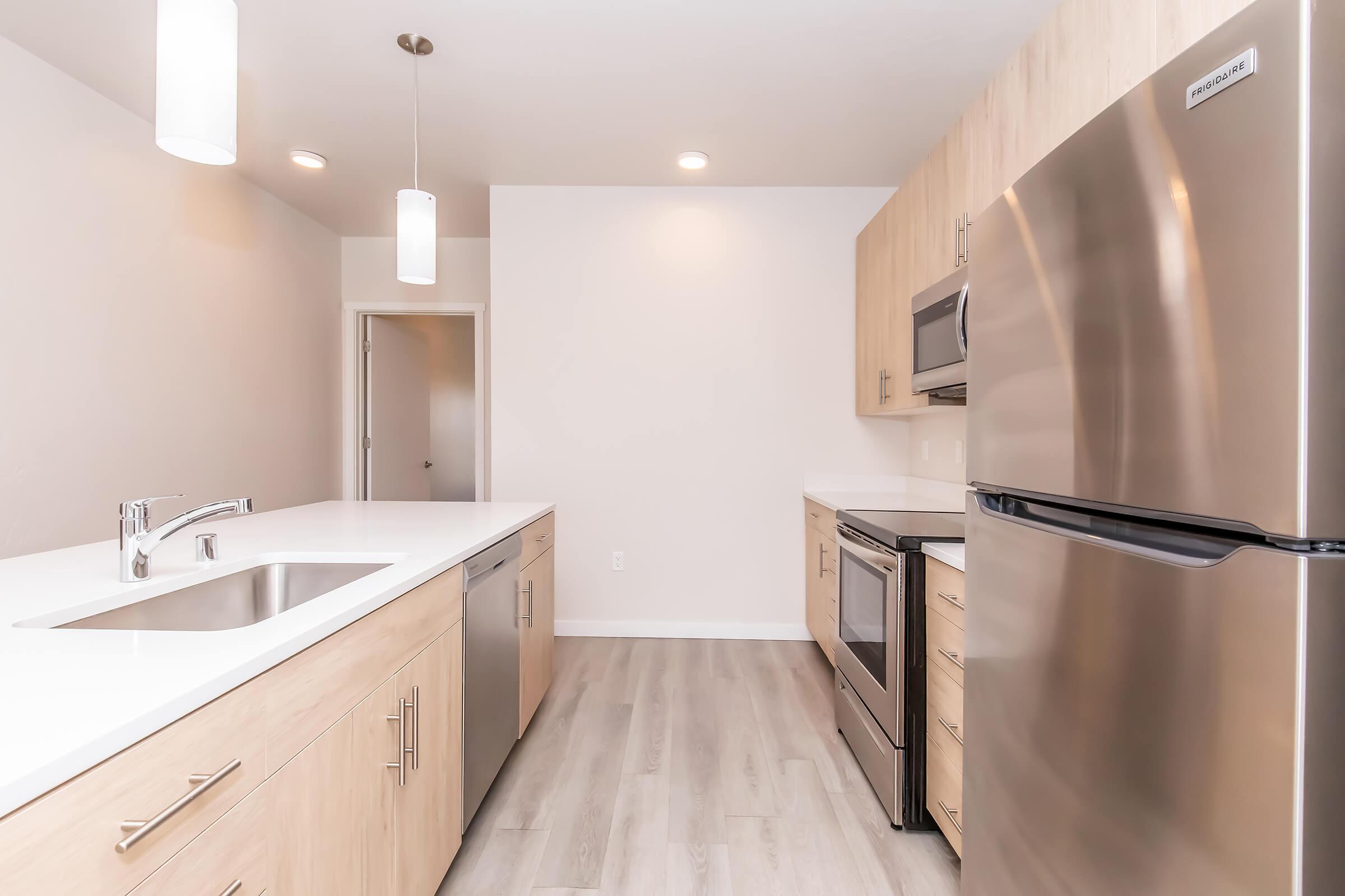 Modern kitchen featuring light wood cabinetry, stainless steel appliances including a refrigerator, oven, and dishwasher, white countertops, and pendant lighting. The space is bright and minimalist with a light-colored floor and an open layout.