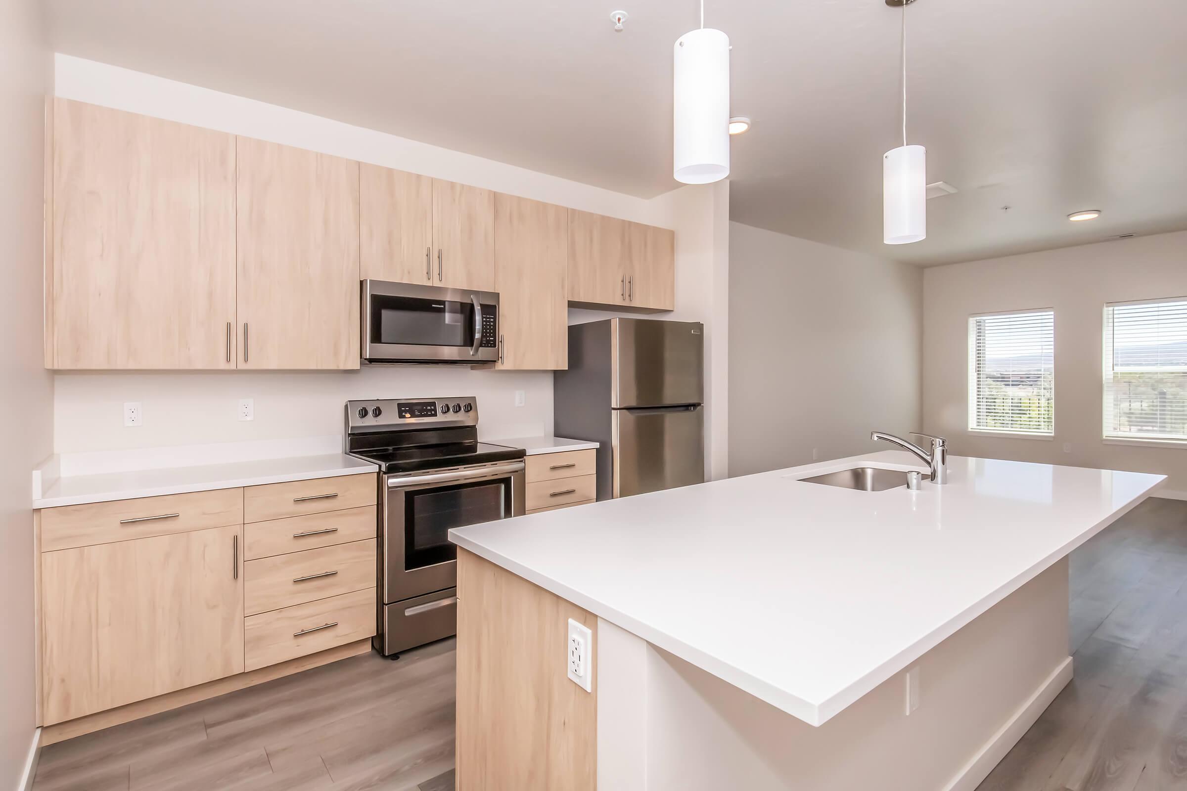 Modern kitchen featuring light wood cabinetry, stainless steel appliances including a microwave and oven, and a large white countertop island. The space has large windows allowing natural light and is designed with a clean, contemporary aesthetic.