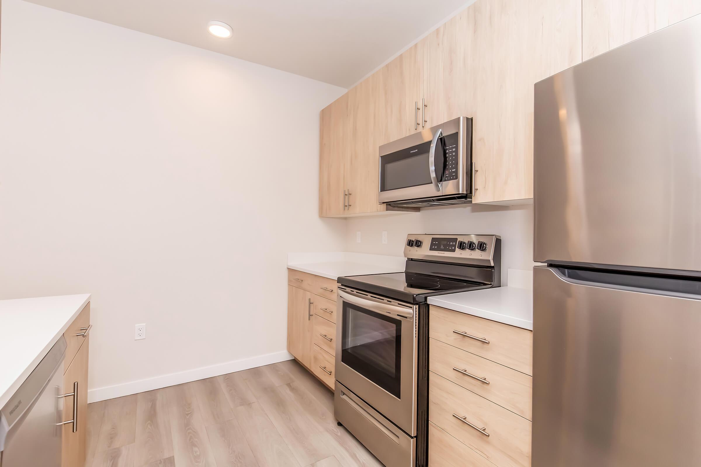 Modern kitchen with light wood cabinetry, stainless steel appliances including an oven, microwave, and refrigerator, and a white countertop. The space features a light, neutral color palette and sleek design, creating a clean and inviting atmosphere. Bright lighting enhances the openness of the area.