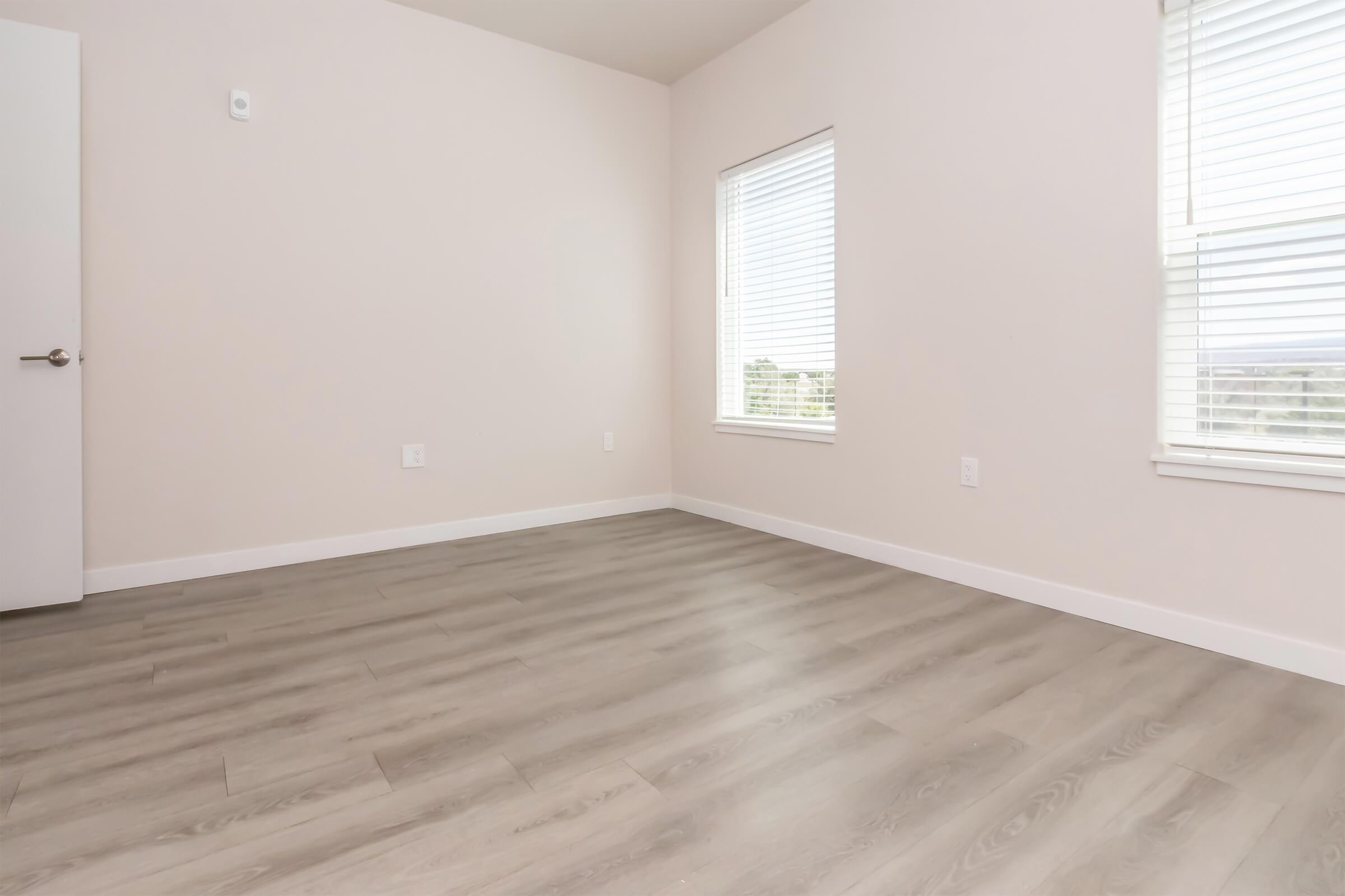 A spacious, empty room with light gray hardwood flooring and pale walls. There are two windows with white blinds allowing natural light to enter, and a white door on the left side. The overall atmosphere is bright and minimalistic, suitable for a variety of uses.