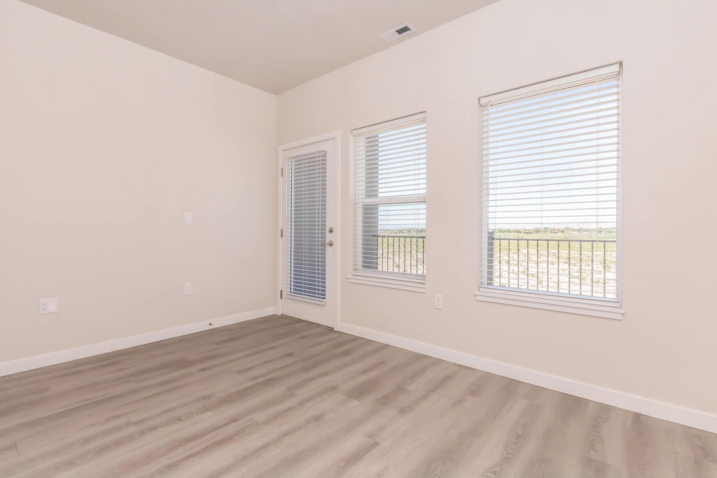A bright, empty room with light-colored walls and large windows featuring blinds. The floor is made of light wood laminate. A glass door leads to an outdoor area, and there is a scenic view outside. The space is spacious and uncluttered, ideal for various furniture arrangements.