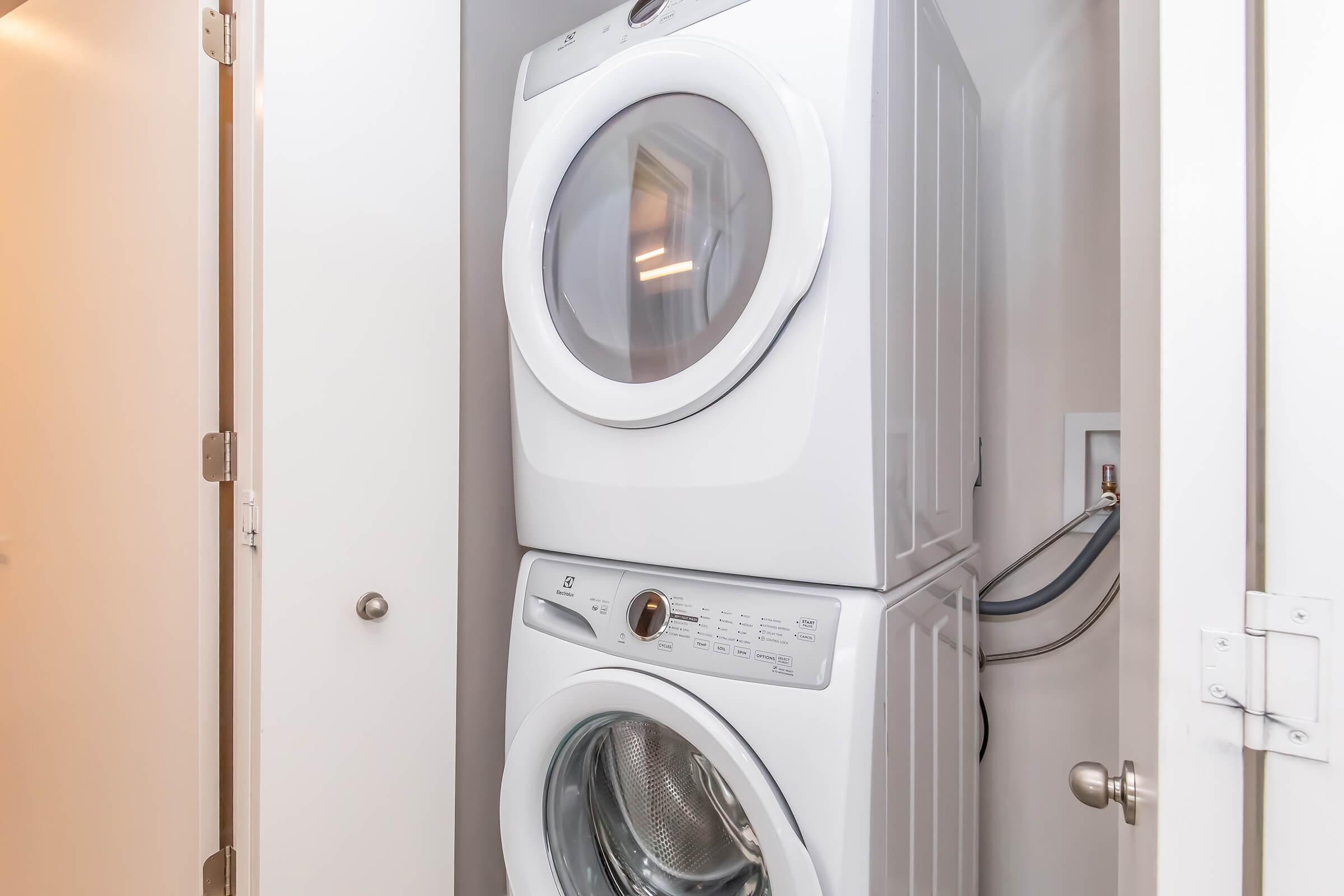 A stacked washer and dryer unit in a compact laundry space. The washer is on the bottom and the dryer is on the top, both in a modern white finish with front-facing doors. The unit is located in a light-colored closet with closed doors and a silver doorknob visible.