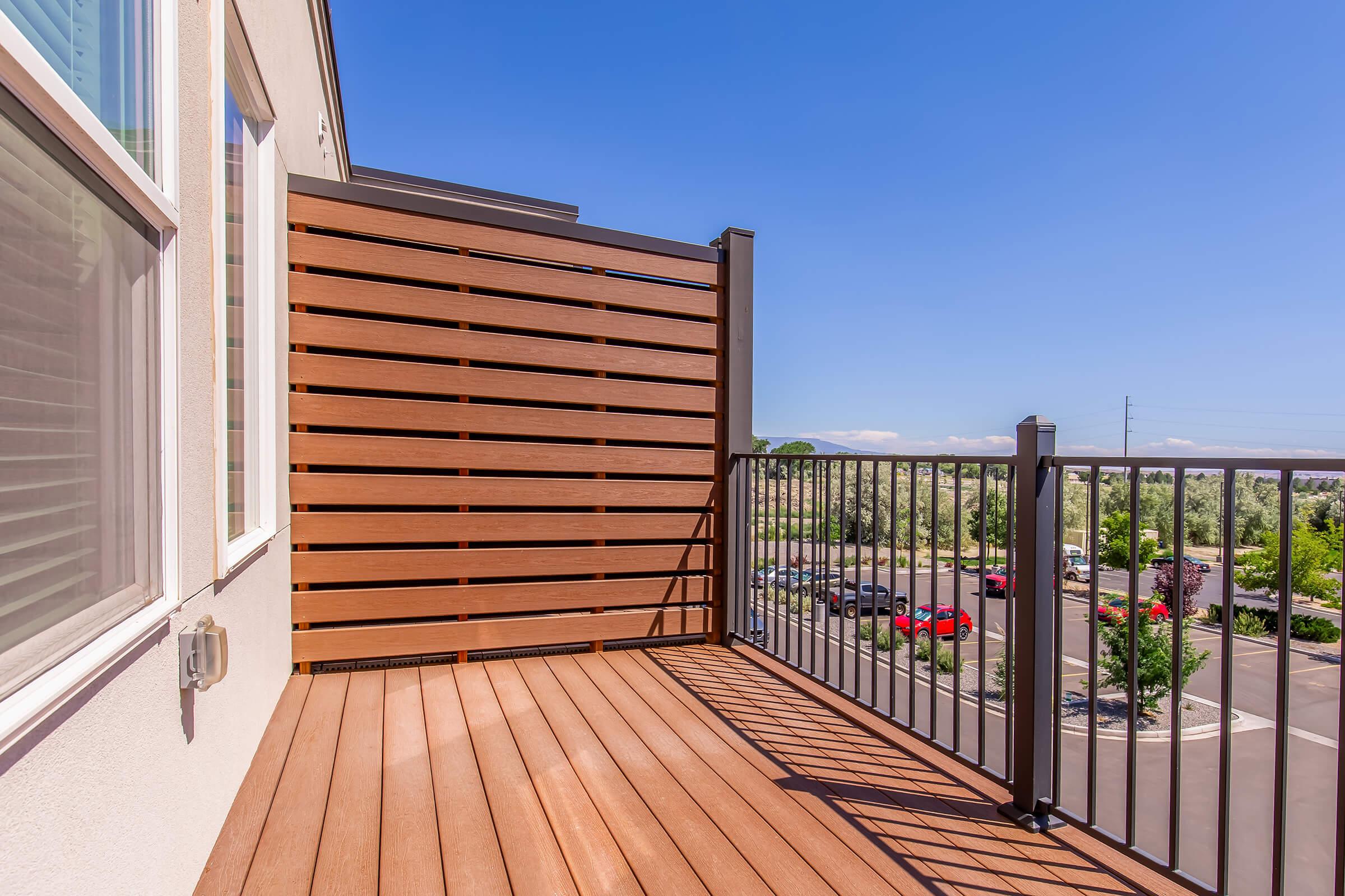 A balcony featuring wooden slat privacy screens and metal railings, with a clear blue sky above. The view includes a landscaped area and parked cars in the distance, suggesting a modern residential environment. Sunlight casts shadows on the wooden deck.